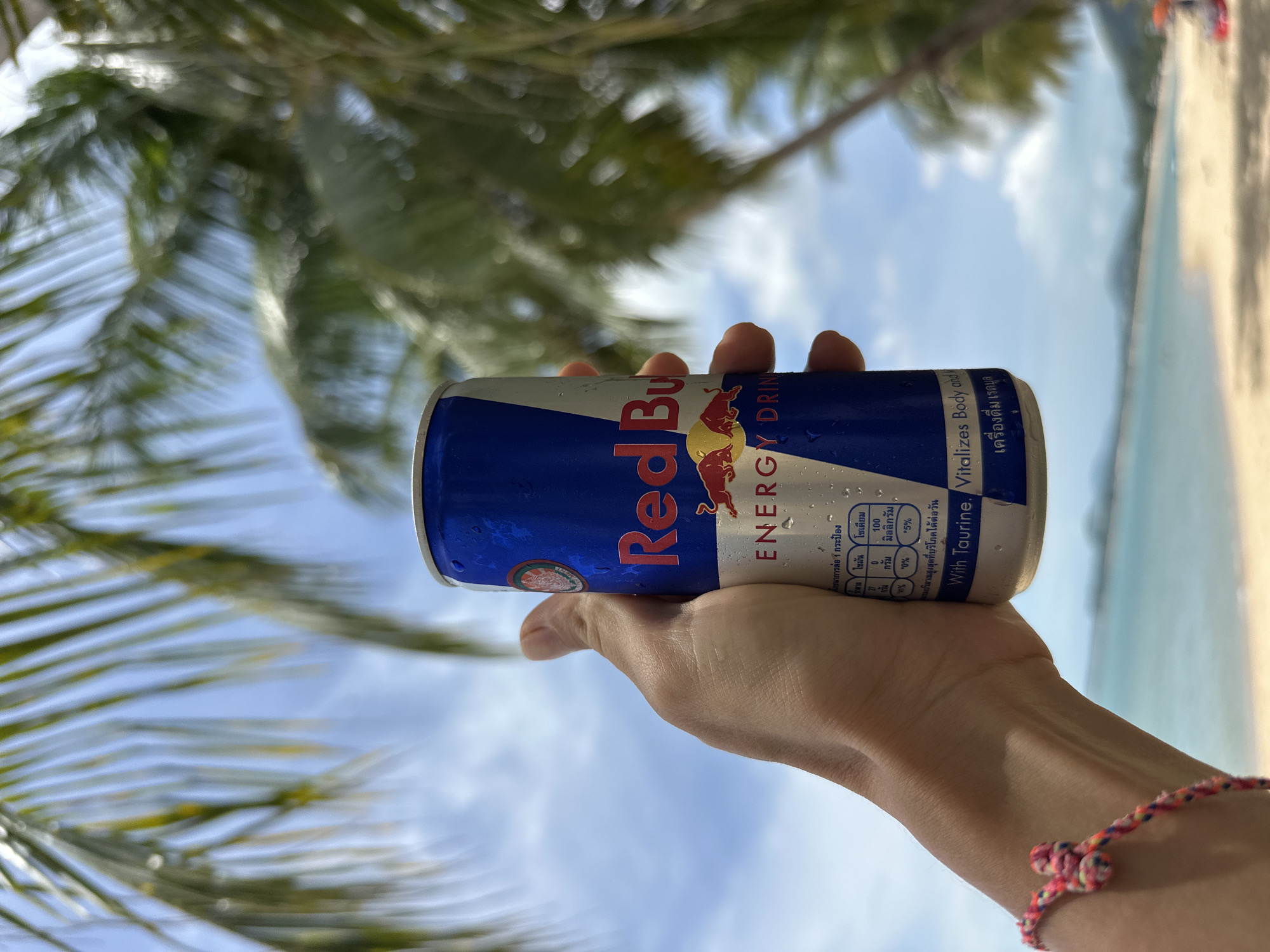 A person's hand holds a can of Red Bull energy drink against a backdrop of palm trees, a beach, and the ocean. The can is blue and white with the Red Bull logo and the words "ENERGY DRINK" visible. There are water droplets on the can, suggesting it is cold. The background is slightly blurred, but a tropical beach scene is discernible with clear blue skies, white sand, and turquoise water. The location is identified as Baan Mae Nam, Thailand, a district on the island of Koh Samui known for its beaches.