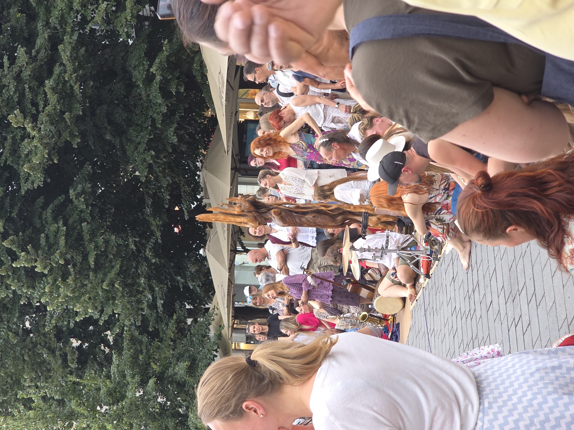 A medium shot captures an outdoor gathering of people in an urban setting, likely a plaza or street, with trees and buildings in the background. The foreground is dominated by a woman wearing a white t-shirt and a blue and white patterned skirt, with her back to the camera. To her right, a group of musicians and spectators are visible. A woman with bright red hair is playing a drum, while a saxophone and other percussion instruments are arranged nearby. Several people are observing the performance, some with engaged expressions. The ground is paved with cobblestones. The overall atmosphere appears to be lively and social, possibly a street performance or a casual outdoor event.