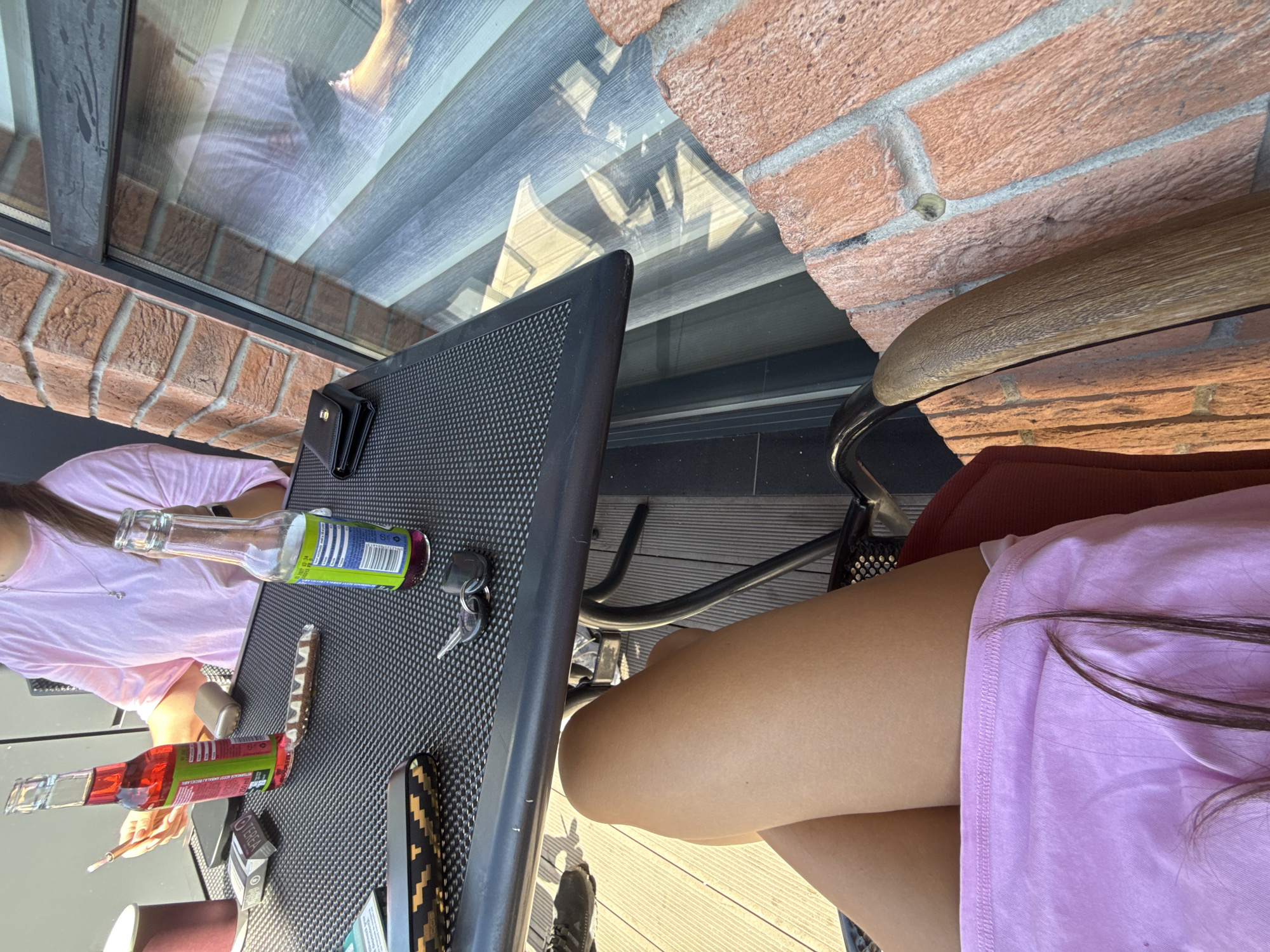 The image depicts a table with two bottles of beverages and other items on it, viewed from a seated perspective. In the foreground, a person's bare legs are visible, indicating they are wearing a light pink garment. The table is dark and textured, with a cigarette pack, a cigarette lighter, a wallet, and a set of keys on its surface. A bottle with a red liquid and another with a purple liquid are present, along with what appears to be a snack bar. The background shows a brick wall and a window with blinds, suggesting an outdoor or semi-outdoor setting. The overall impression is one of a relaxed moment, possibly on a balcony or patio. The scene is in the city of Comrat, Moldova.
