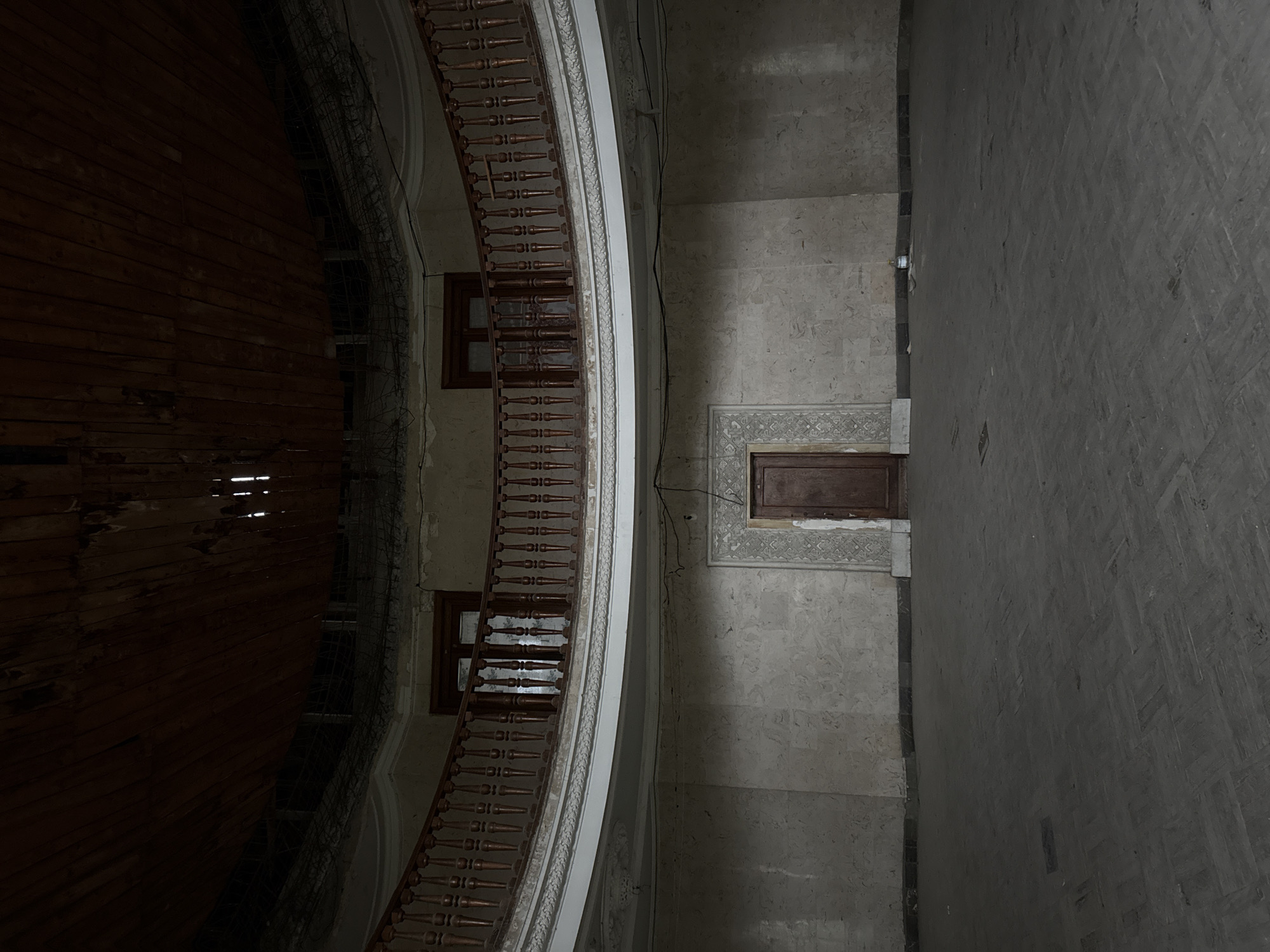 The image depicts an interior view of a multi-story structure. The left side is dominated by a dark, possibly wooden, surface with some visible light gaps. Along the center, a curved, wooden railing runs alongside the wall, with two windows visible behind it. The right side of the image showcases a gray stone wall, featuring a door with decorative trim above it. The lighting appears dim and uneven, suggesting an older, possibly abandoned or neglected building. No people are visible. The image does not contain enough information to definitively confirm the location in Tskaltubo, Georgia, but the architectural style is consistent with buildings in that region.