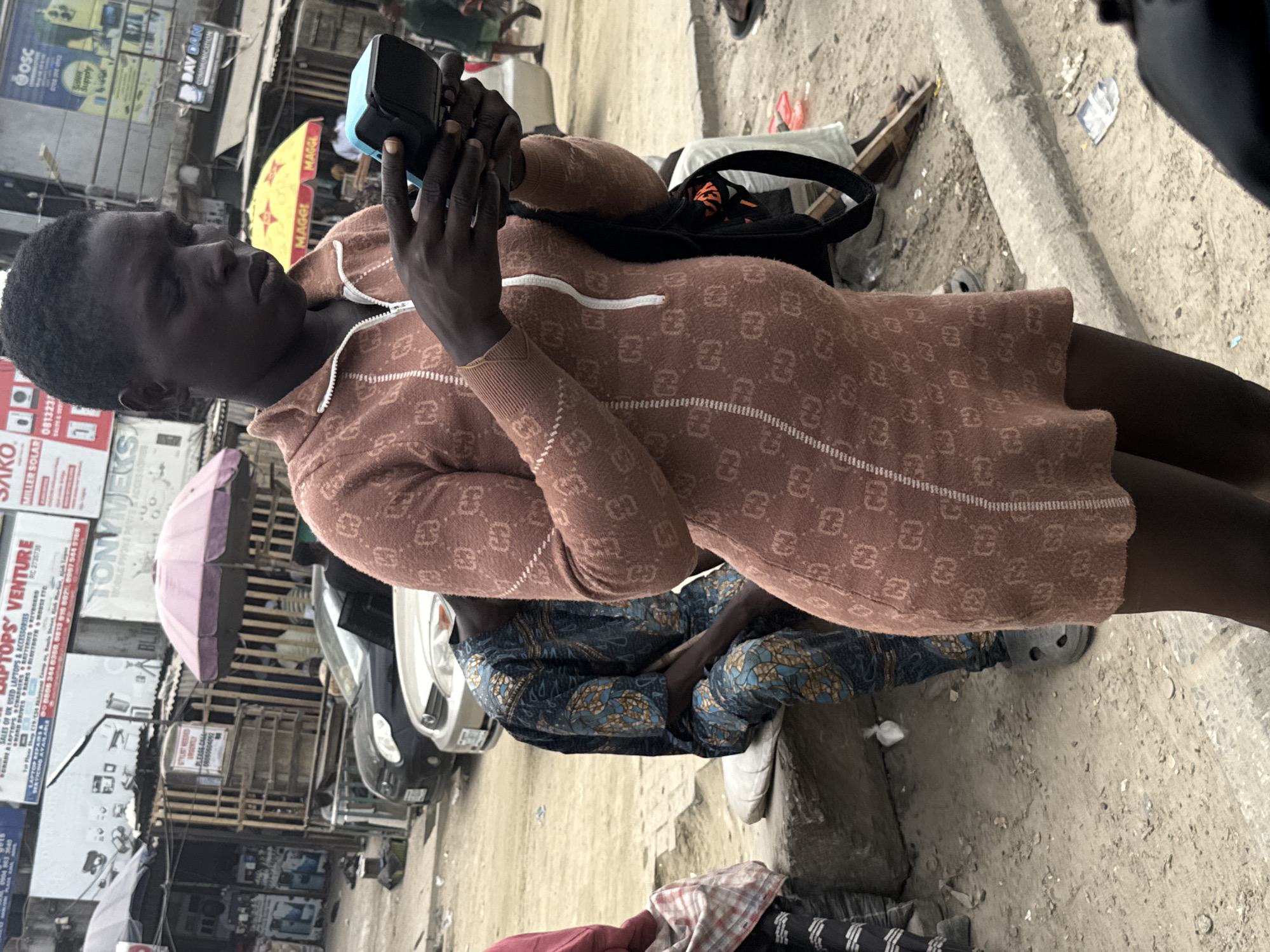 In Lekki, Nigeria, a woman stands on a dirt road, her hands engaged with a mobile point-of-sale device. She is wearing a brown sweater dress with a repeating pattern of interlocking 'G' logos. To her left, a person in a blue and gold patterned traditional garment sits on a concrete block, facing away from the camera. In the background, a parked silver car and a black car are visible. Above them, a collection of buildings and signs, including one that reads "TONYMJEY MOBILE PHONE PARTS & ACCESSORIES," suggests a commercial area. The environment is dusty, with scattered debris on the ground. Two open umbrellas, one pink and one yellow, are also present.