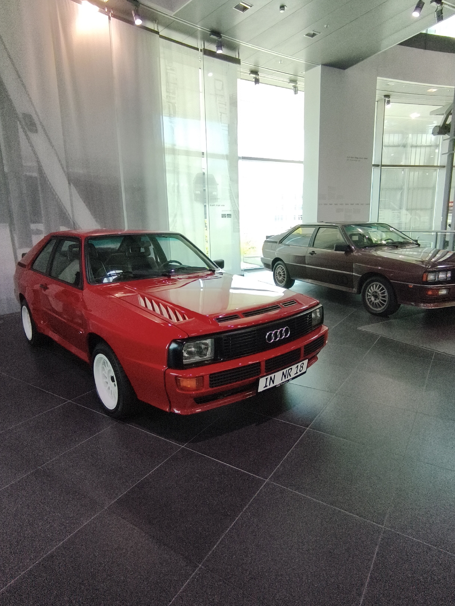 In Ingolstadt, Germany, two Audi Quattro sports cars are displayed indoors on a dark tiled floor. The car in the foreground is a bright red, two-door coupe with white rims, its front license plate reading "IN NR 18." To the right and slightly behind is a darker colored, possibly brown, Audi Quattro of a similar model. The cars are presented in what appears to be a museum or exhibition space, with large windows and sheer white curtains in the background. The lighting suggests an indoor setting with overhead illumination. There are no people visible in the frame.