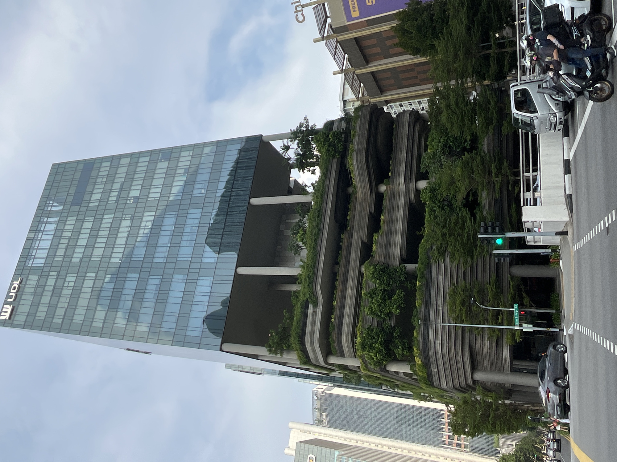 The image depicts a street scene in Singapore, featuring a prominent glass skyscraper with a unique terraced design covered in lush greenery. The skyscraper has the "UOL" logo visible at the top. In the foreground, a gray sedan is turning at an intersection. A motorcycle carrying two people, both wearing helmets and face masks, is also present in the intersection. Further down the street, a white delivery truck and other vehicles are visible. Traffic lights and street signs, including one that reads "New Bridge Rd," are present. The sky is partly cloudy.