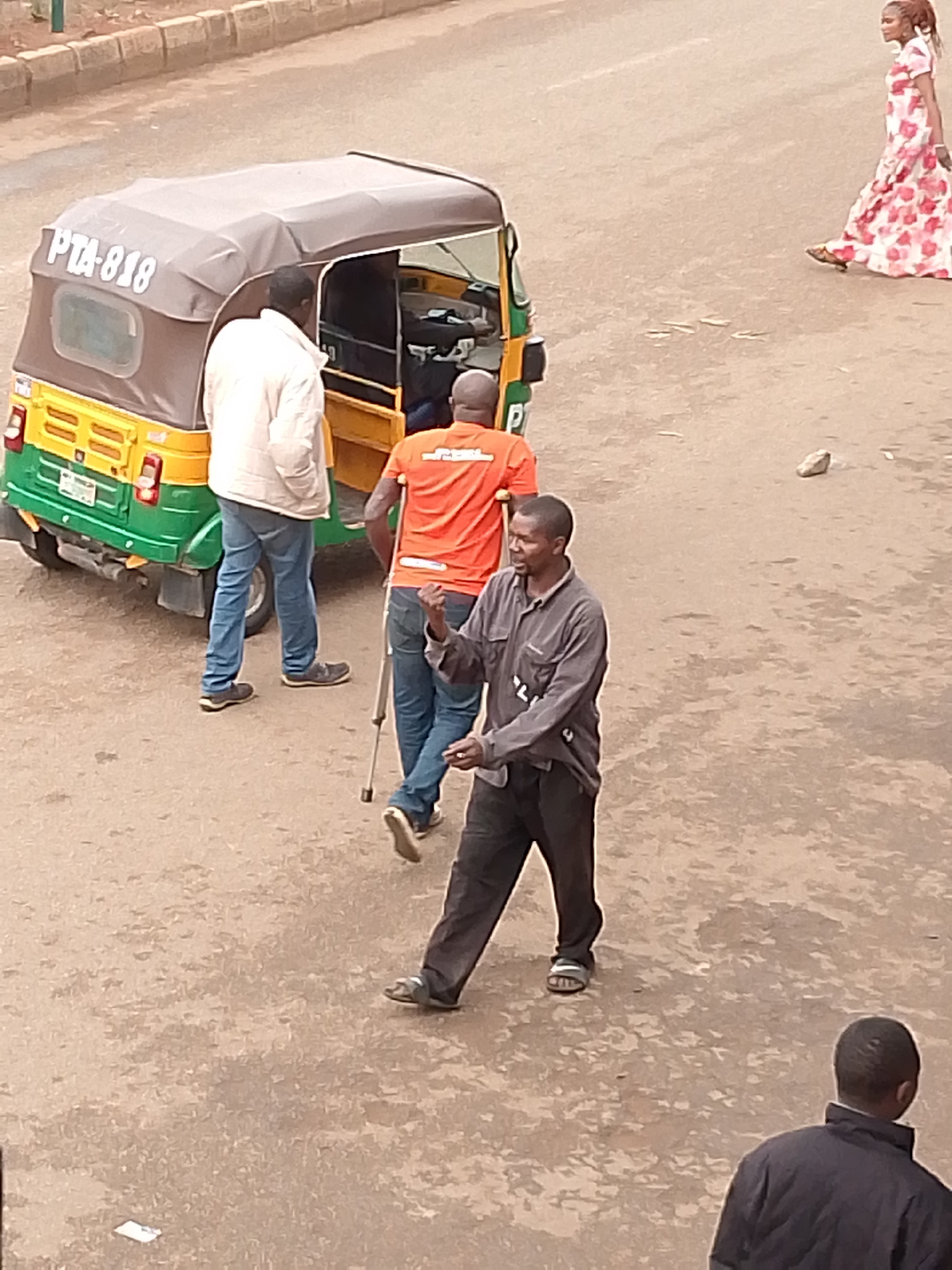 The image depicts a street scene in Bukuru, Nigeria. A three-wheeled motorized vehicle, numbered PTA-818, is visible on the road. Four people are present: one in a white jacket and blue jeans, another in an orange shirt and blue jeans using a crutch, one in a gray shirt and black pants wearing sandals, and another in a black jacket. A woman in a long floral dress is walking in the background. The surface of the road appears worn.