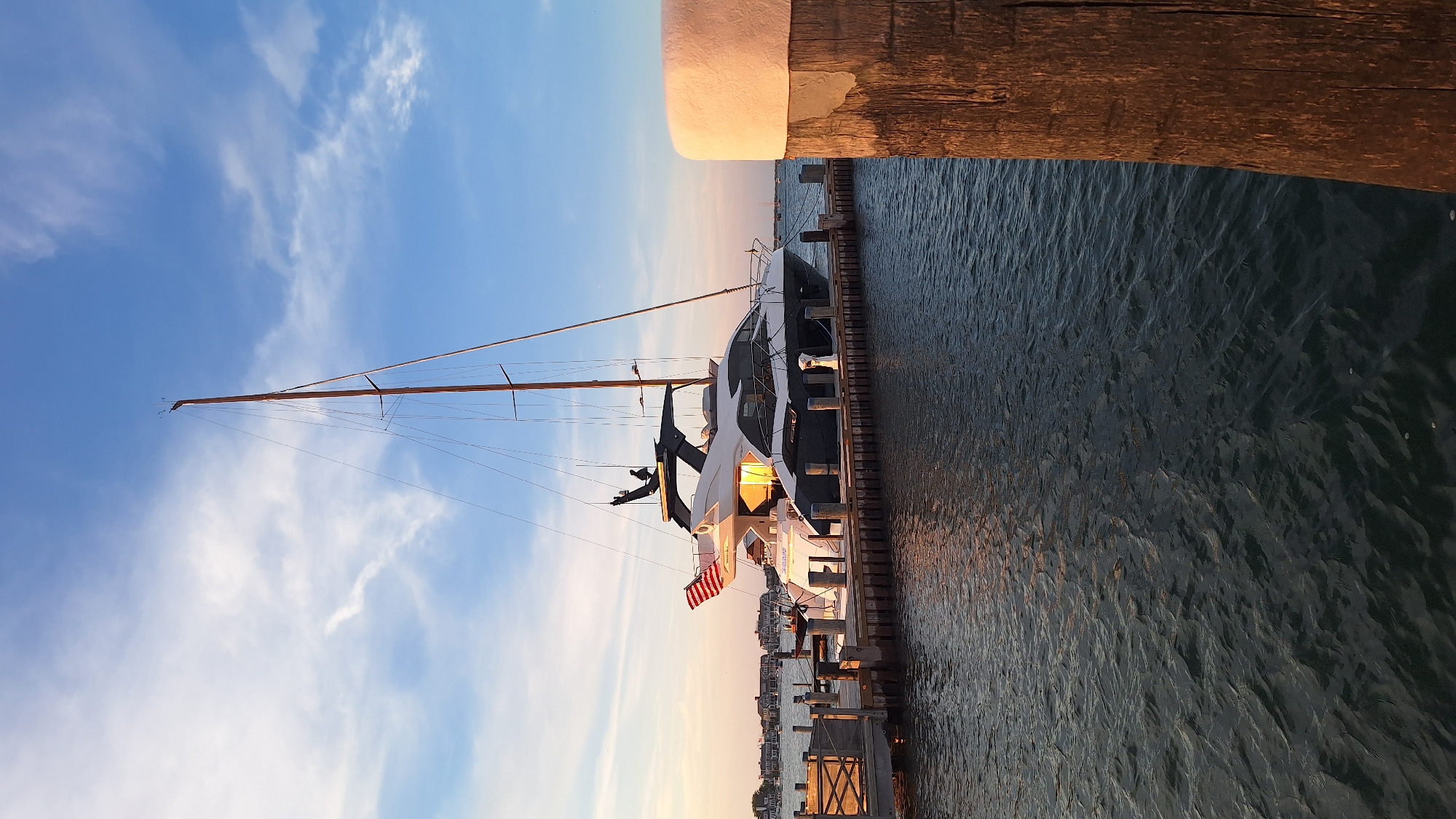 A large, modern yacht with a dark hull and white superstructure is docked at a wooden pier. The yacht has a tall mast and is flying an American flag. The scene appears to be during sunset, with warm light illuminating the yacht and casting reflections on the water. In the background, there are houses and other boats, suggesting a harbor or marina. The water is calm with gentle ripples. The sky is a mix of blue and orange, with scattered clouds. A weathered wooden post with a light-colored top is in the foreground on the right side of the image. The overall setting is likely a coastal town, possibly Nantucket, USA, given the architectural style of the houses in the background and the presence of a yacht.