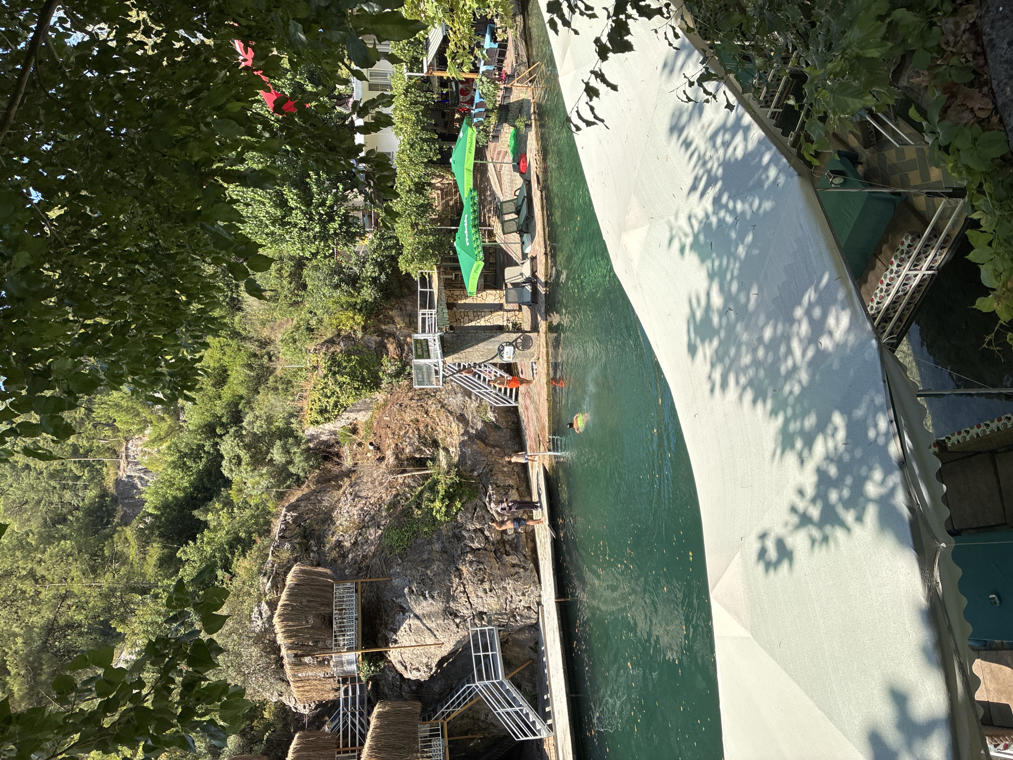 This image depicts a serene outdoor setting, likely a natural pool or river, surrounded by lush greenery and rocky cliffs. The scene is vibrant, with sunlight filtering through the trees, casting dappled shadows on the water and surrounding structures.
In the foreground, a large, light-colored canopy stretches across the frame, its white fabric illuminated by the sun. Shadows of leaves and branches dance on its surface, suggesting the presence of overhead trees. Beneath the canopy, a glimpse of tables and seating hints at a dining or resting area.
The main focus of the image is the body of water, a deep green, calm pool. Several people are visible in and around the water. In the middle ground, a person is swimming, their head visible above the surface. On a wooden platform bordering the water, several individuals are standing. One person, wearing a black headscarf and a long, dark dress, stands with another person in casual attire. To their right, a child in a blue swimsuit stands near the water's edge. Further to the right, on another section of the platform, a person in an orange swimming trunk is visible near a set of stairs.
The background features a steep, rocky cliff face. Several multi-level structures with thatched roofs are built into the cliffside, accessed by white staircases. These appear to be unique accommodations or resting spots. The cliff itself is covered in dense vegetation, with trees and bushes clinging to its surface.
Further into the background, a white building with windows is visible, nestled amongst the trees. In front of the building, two large green umbrellas provide shade over lounge chairs, suggesting an area for relaxation. Tables are also visible, set up for dining, with blue tablecloths.
The overall impression is a tranquil retreat, blending natural beauty with comfortable amenities. The location is identified as Alanya, Turkey, a popular coastal city known for its historical sites and natural attractions. The scene likely represents a resort, a restaurant with a natural pool, or a recreational area within Alanya.