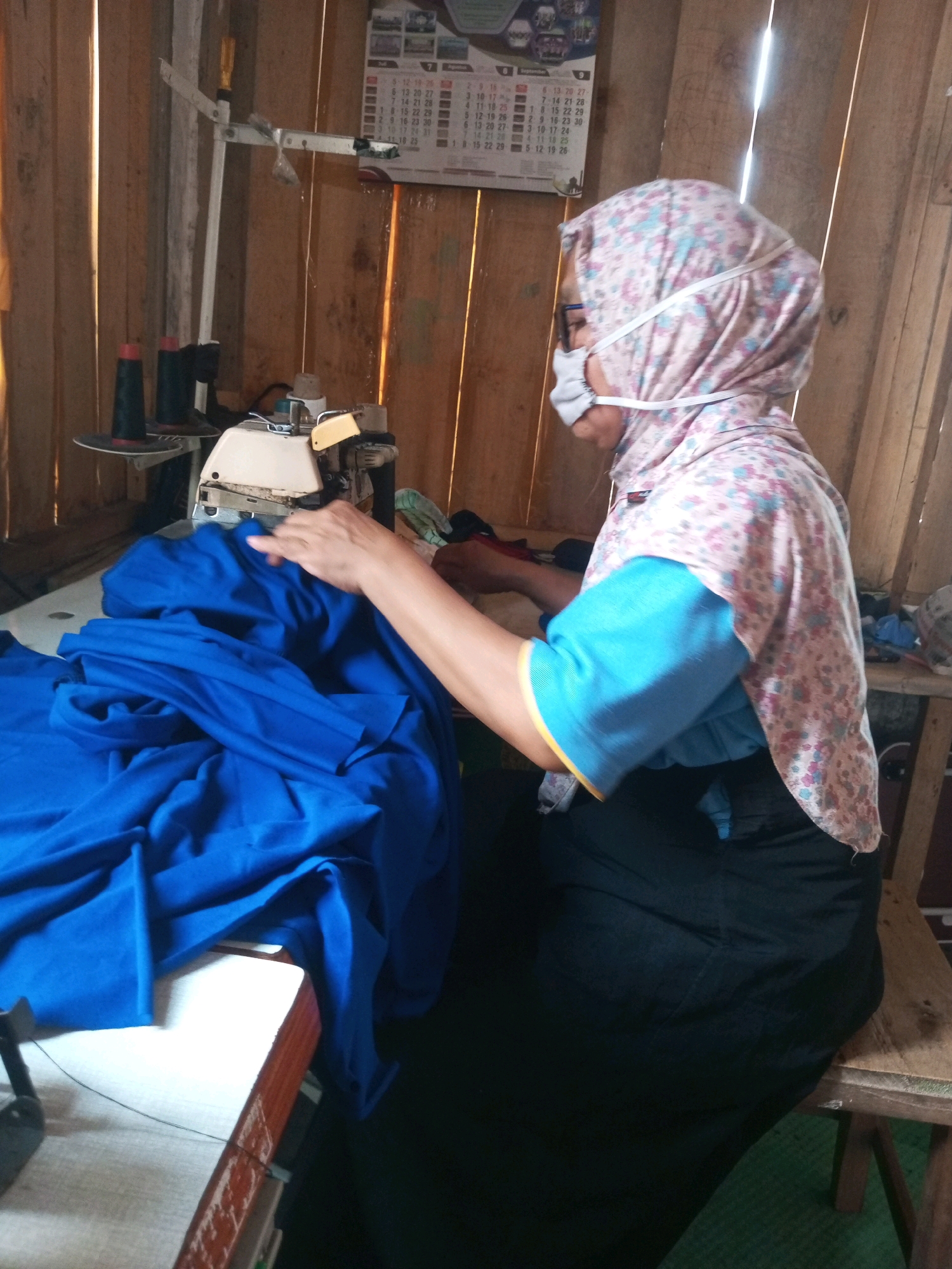 A person, wearing a pink floral headscarf, a blue t-shirt with yellow trim, and a dark skirt, sits in front of a sewing machine. The person is also wearing a white face mask and glasses, with the mask secured by elastic straps that go around the headscarf. The person's right hand is near the sewing machine, and a large amount of bright blue fabric is piled on the table in front of the machine. The background consists of vertical wooden planks and a calendar hanging on the wall. A spool of thread is visible on a stand to the left of the sewing machine. The scene appears to be an indoor workspace, possibly a home or small workshop, in Kalibeber, Indonesia.