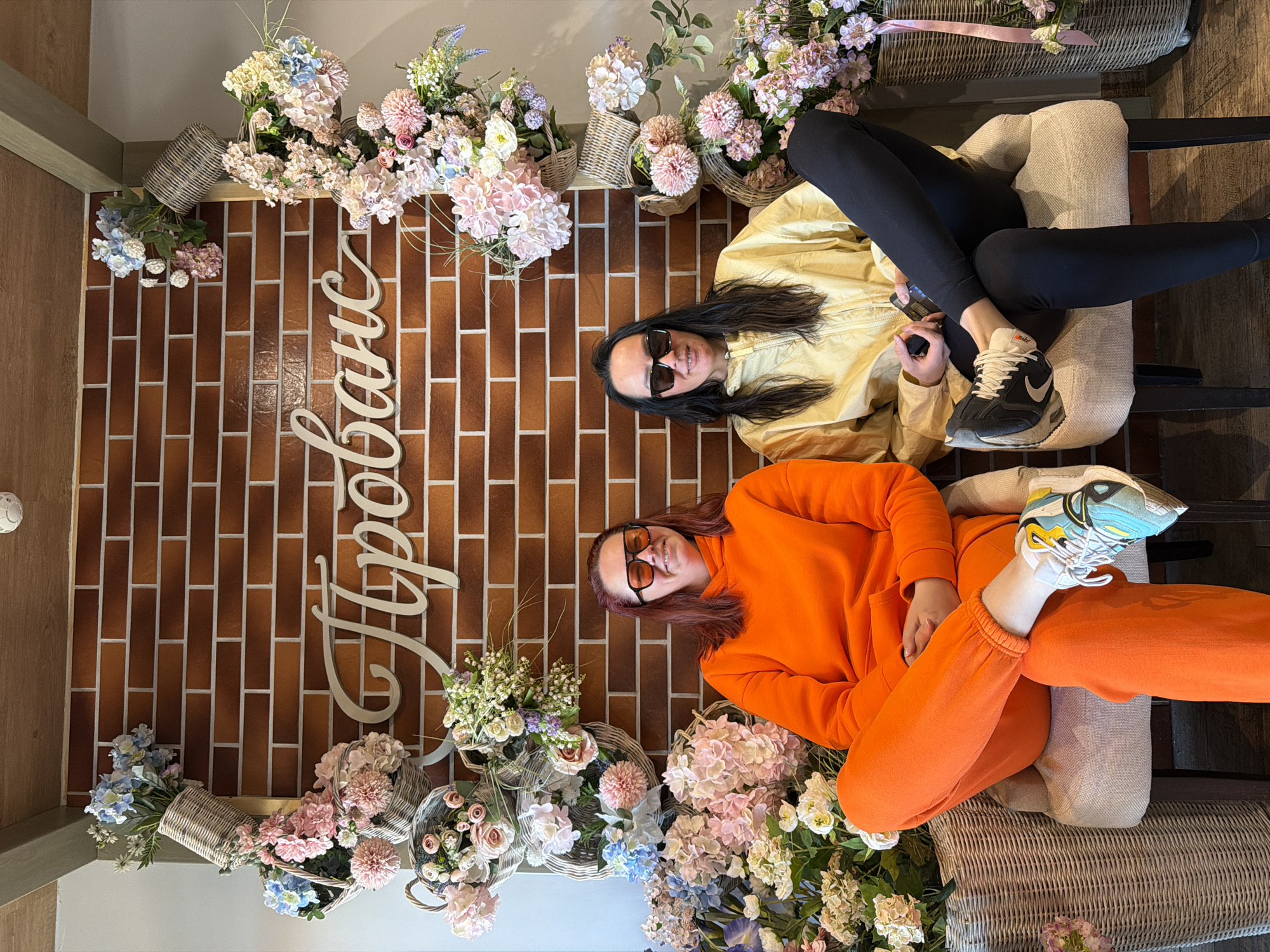 Two women are sitting on wicker chairs in front of a brick wall decorated with baskets of flowers and a white cursive sign. The woman on the left wears an orange tracksuit and orange-tinted sunglasses. The woman on the right wears black leggings and a yellow jacket with black sunglasses, and holds a black object, possibly a phone or wallet, in her hand. The location may be Votkinsk, Russia, due to the Cyrillic script on the sign.