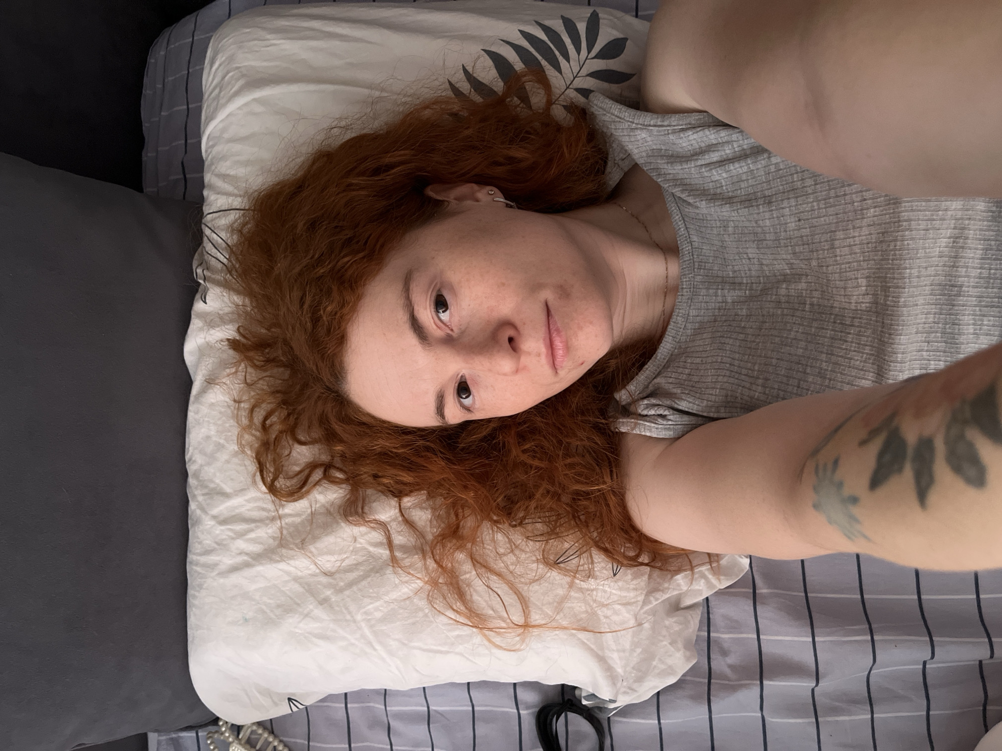 A person with long, wavy red hair and freckles is lying on their back on a bed, looking directly at the camera. The image is captured from an overhead perspective, likely a selfie. The individual wears a light grey, ribbed t-shirt and has a small silver stud earring in their right ear. A thin, gold-colored chain necklace is visible at the collarbone. A tattoo, depicting what appears to be a stylized floral or leafy design in muted blues and greys, is visible on their upper left arm.
The person's voluminous red hair is spread across a white pillow decorated with a dark grey leaf pattern. The bed is covered with grey and white striped sheets. A dark grey pillow or blanket is positioned to the left of the main pillow. In the bottom right corner of the frame, a black hair tie or similar accessory is partially visible. The lighting is soft and even, suggesting an indoor setting, possibly a bedroom, during the daytime. The person exhibits a neutral to slightly contemplative expression. No text is visible in the image. The location context is Chaikovsky, Russia.