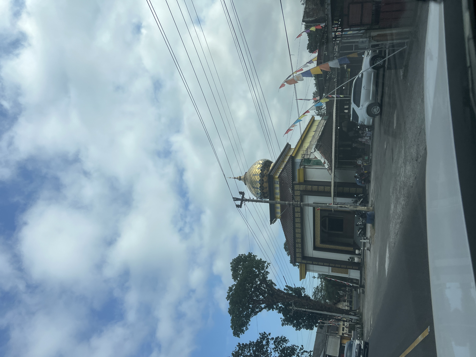 The image shows a street scene in Badran, Indonesia, with a mosque as the prominent structure. The mosque features a golden dome and is adorned with decorative gold and white patterns. In front of the mosque, there is a sheltered area with seating where several people are gathered. Several motorcycles are parked near the entrance of the mosque. To the left of the mosque, a large, distinctive tree with a heart-shaped canopy stands tall. A silver SUV is parked in front of a building to the right of the mosque. Colorful flags are strung across the scene, indicating a possible celebration or event. The sky is partly cloudy, with a mix of blue sky and white clouds. The road in the foreground appears to be asphalt, with a yellow line marking the lane. The image is taken from inside a vehicle, as suggested by the reflection on the windshield in the foreground.