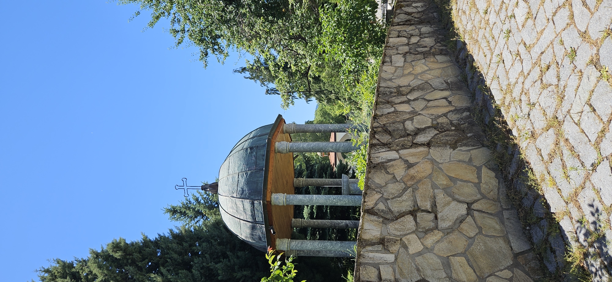 The image depicts a rotunda with a domed, green-patinated metal roof topped with a cross, supported by marble-like columns. A dry-stone wall constructed from irregular, light-colored stones forms a barrier in the foreground, with a paved pathway to its right, speckled with sparse vegetation. Behind the wall, lush green trees, including conifers, surround the rotunda, and a clear blue sky dominates the upper portion of the frame. In the distance, beyond the trees, a glimpse of another building with a red roof and white walls can be seen. The scene appears to be outdoors in a serene, possibly religious or memorial setting. The location is identified as Ovchartsi, Bulgaria. There are no people or active interactions visible in the image.