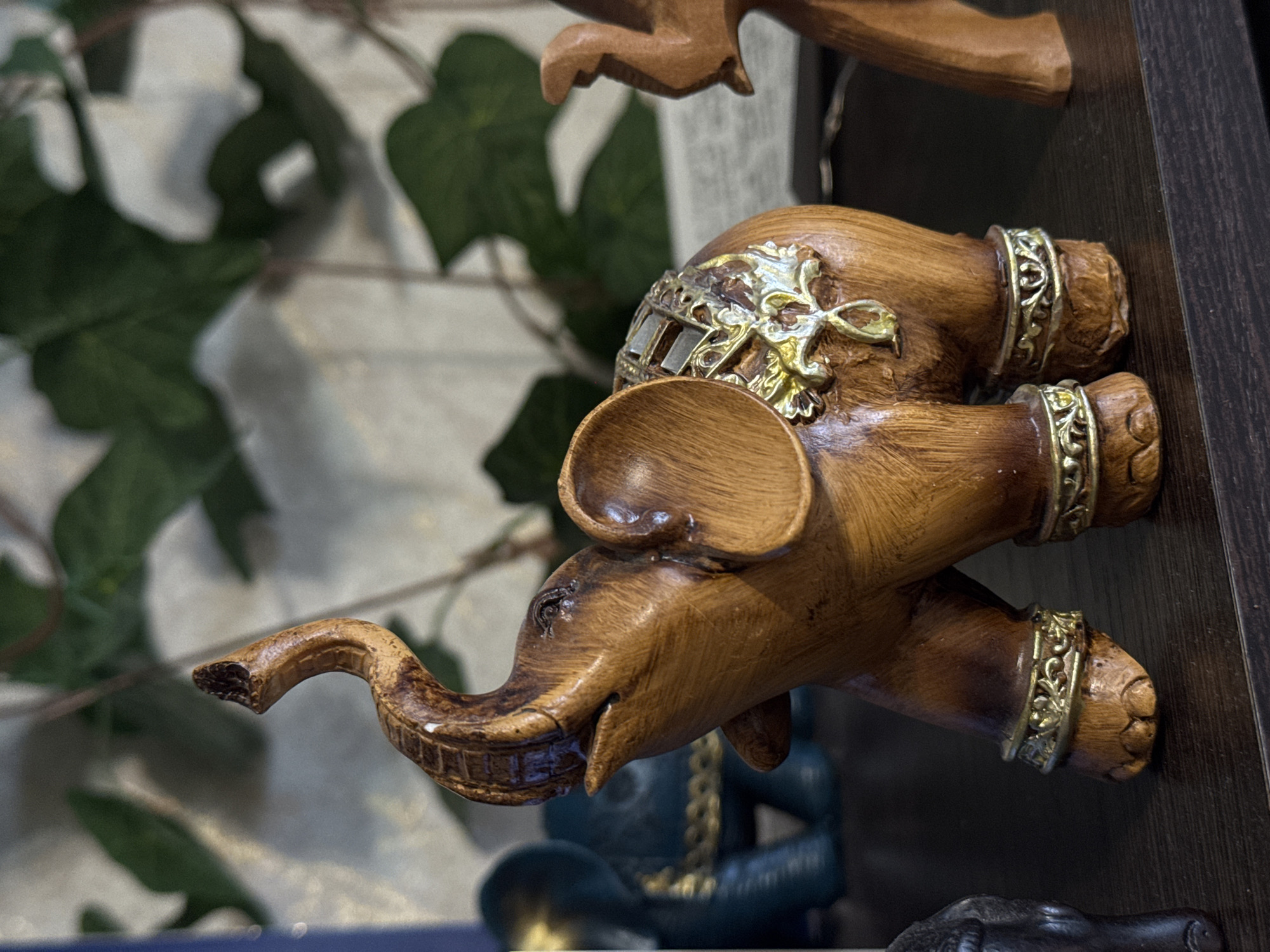 The image captures a decorative wooden elephant figurine, oriented horizontally across the frame, displayed on a dark brown wooden shelf or surface. The elephant is intricately carved from a medium-brown wood with visible grain and a polished appearance. It is adorned with ornate gold-colored metallic embellishments. A large, detailed golden decoration, possibly representing a saddle or ceremonial blanket, featuring mirrored or textured segments and intricate floral patterns, rests on its back. Smaller golden bands with similar decorative motifs encircle its legs, resembling anklets. The elephant's trunk is raised and curved upwards, showcasing carved details, and its large ears are prominent. Its eyes are small and dark.
The background is softly out of focus, revealing blurred green leaves, likely from an indoor plant, against a lighter, indistinct surface. Below the main elephant figurine, hints of other decorative items are visible. These include a dark blue or green figurine, possibly a Buddha statue, with faint golden chains around its neck or chest, and another dark, amorphous carved object at the very bottom of the frame. The lighting suggests an indoor setting. There is no discernible activity or visible text in the image.