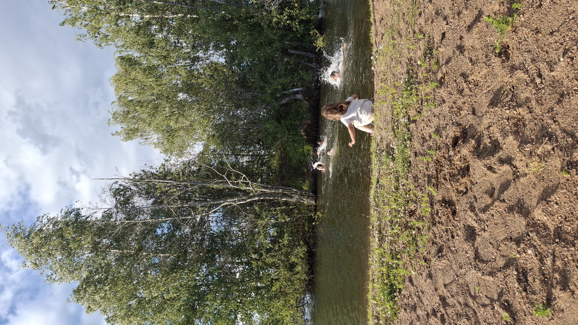 The image depicts a group of people swimming and splashing in a body of water, likely a lake or river, surrounded by lush green trees. In the foreground, a young child with brown hair, wearing a white t-shirt and shorts, sits on a sandy bank, facing the water. The child's left hand is extended towards the water. Several individuals are visible in the water, some partially submerged and creating splashes, indicating active swimming or playful activity. The water is calm except for the areas where the people are swimming. The background is dominated by a dense line of trees with green foliage, under a partially cloudy sky. The sandy bank in the foreground shows footprints and some sparse green vegetation. The location is identified as Innolovo, Russia.