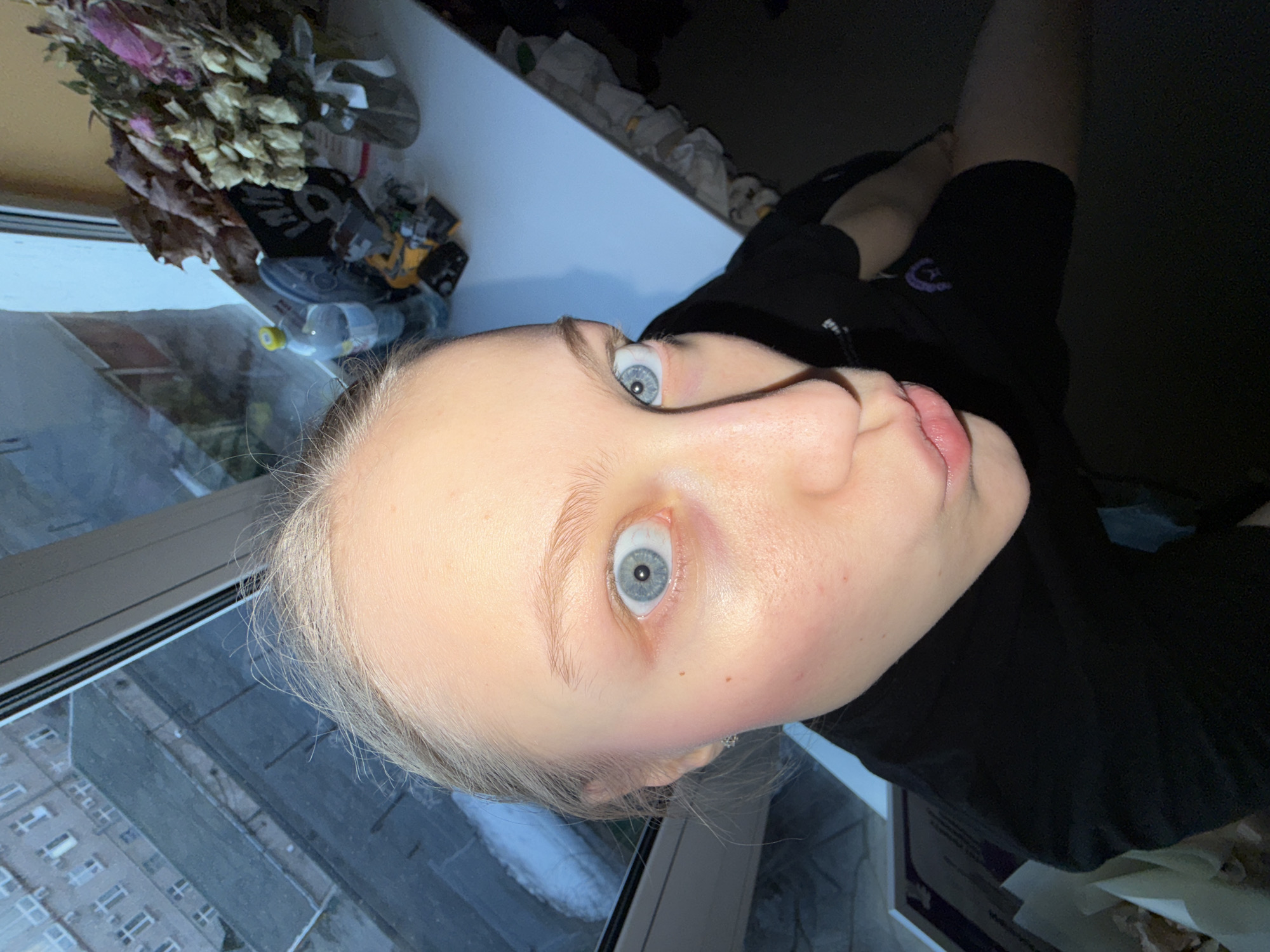 A selfie taken indoors, likely in Perm, Russia, based on the building visible through the window. The image captures a person with light skin, pale hair, and blue eyes looking directly at the camera with a slight upward angle. They are wearing a black top. The background features a window with a view of an urban environment, including a multi-story building with numerous windows, suggesting an apartment complex. There is a bouquet of dried flowers or plants on the windowsill. The lighting suggests it is daytime. The image includes trash and other items on the windowsill.