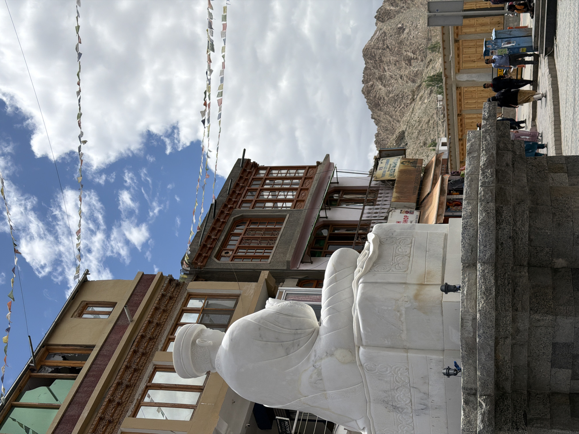 This is a medium shot, eye-level, outdoor photograph taken in the city of Leh, India. In the foreground, a large white marble statue of a seated figure in meditation is positioned on a stone base. The statue is facing away from the camera, with its head turned slightly to the left. The figure wears traditional monastic attire. The base of the statue is intricately carved with decorative patterns. Two small, black spotlights are mounted on the front of the stone base, pointing upwards.
Behind the statue, a multi-story building with a yellowish facade features several windows with wooden frames. Prayer flags are strung across the sky above the building. To the right of this building is another building with a darker facade and wooden-framed windows. Further to the right, a street scene unfolds with several people walking. A blue metal trash bin is visible on the right side of the street. In the background, rugged, mountainous terrain dominates the horizon under a sky filled with scattered clouds. The overall lighting suggests it is daytime.
