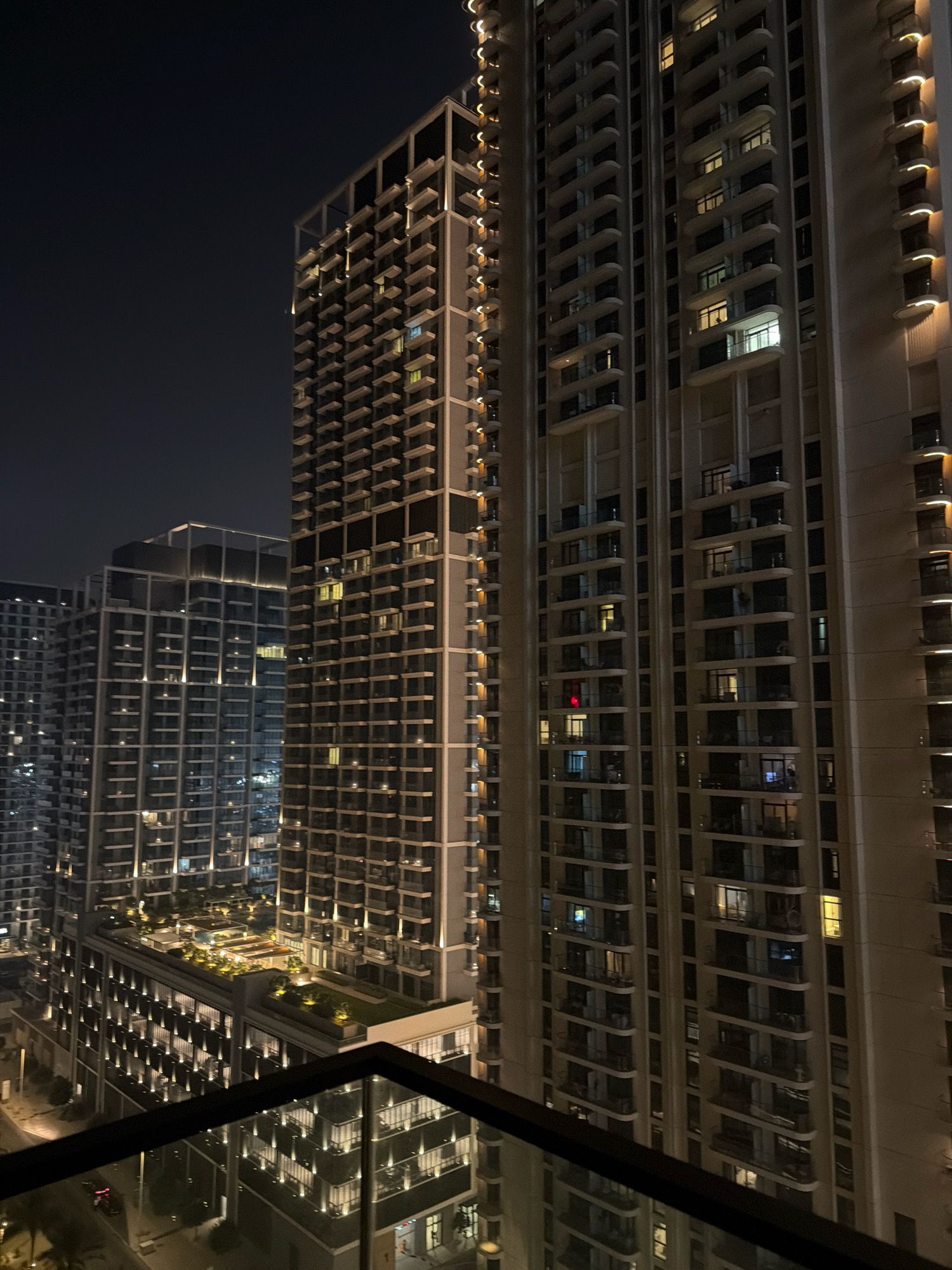 This is a nighttime, high-angle, medium shot of several modern skyscrapers in Dubai, United Arab Emirates. The buildings are tall and narrow, with numerous balconies and windows lit from within. Vertical lighting accents the facades of the buildings, creating a warm glow against the dark night sky. In the foreground, a balcony railing is visible, suggesting the photo was taken from a similar vantage point. One of the buildings in the mid-ground has a green rooftop with strategically placed lighting, possibly indicating a garden or recreational area. The overall impression is one of urban density and architectural modernity, characteristic of Dubai's skyline.