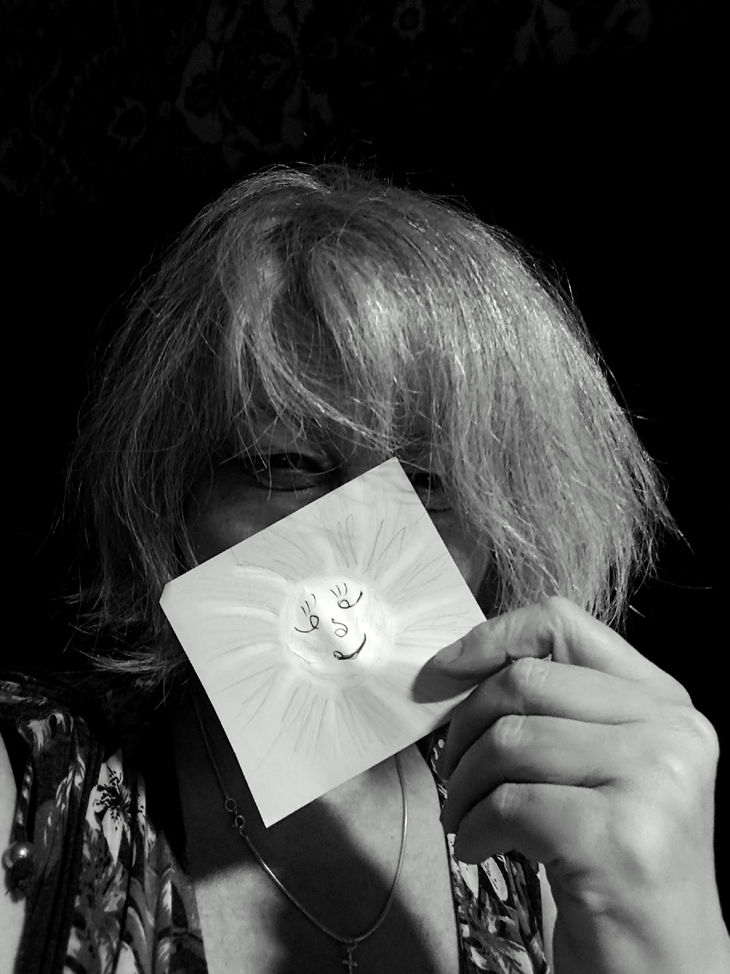 A grayscale, medium shot photograph depicts a person holding a drawing of a smiling sun in front of their face. The person's hair is light-colored and somewhat disheveled, falling around their head. Their eyes are visible behind the drawing, but their expression is obscured. The drawing is on a white, square piece of paper, with simple lines representing the sun's rays and a smiley face. The person is wearing a patterned top and a necklace with a pendant. The background is dark and appears to be a patterned fabric, possibly a curtain or wallpaper. The lighting is dramatic, with the subject's face and the drawing well-lit and the background in shadow. This image is not located in Krasnodar, Russia.