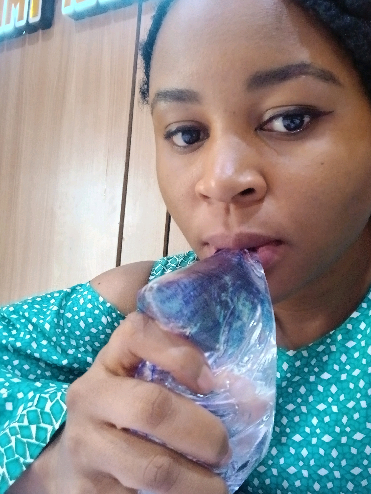 A close-up, waist-up shot captures a person, likely female, holding a blue, transparent plastic bag containing what appears to be ice or a frozen substance to their lips. The individual is wearing a teal-colored top with a white diamond pattern. The background features a light brown, wooden surface and a section of what looks like illuminated signage. The subject's face is partially visible, with attention drawn to their mouth as they interact with the bag. The setting is indoors, indicated by the interior wall and lighting. The image does not provide any specific geographical markers for Gura Top, Nigeria.