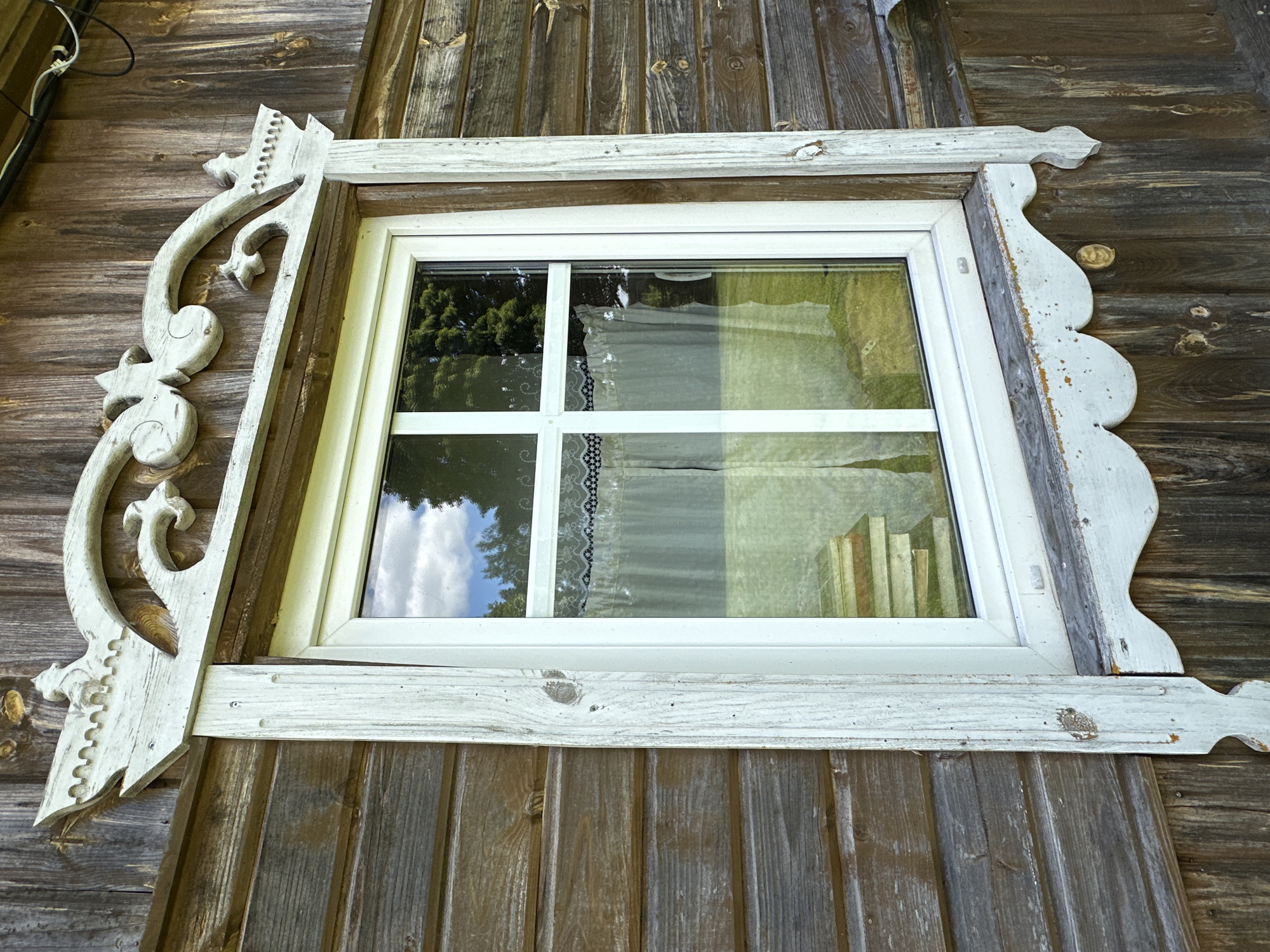 The image shows a close-up of a white-framed window on the exterior of a wooden building in Norviliškės, Lithuania. The window is divided into four panes by white mullions and transoms. The exterior of the building is clad in weathered, horizontal wooden planks. The window is adorned with an ornate, carved wooden trim painted white, featuring decorative elements such as swirls and what appear to be small figures or crests at the top. The trim has a slightly distressed appearance, with some areas of paint worn away. Through the window, a light-colored curtain with a lace trim is visible, and reflections of trees and sky can be seen on the glass. A stack of what appear to be wooden planks is reflected on the lower part of the window. The overall scene suggests a traditional or rustic architectural style, likely part of the Norviliškės Manor complex or a similar historical structure.