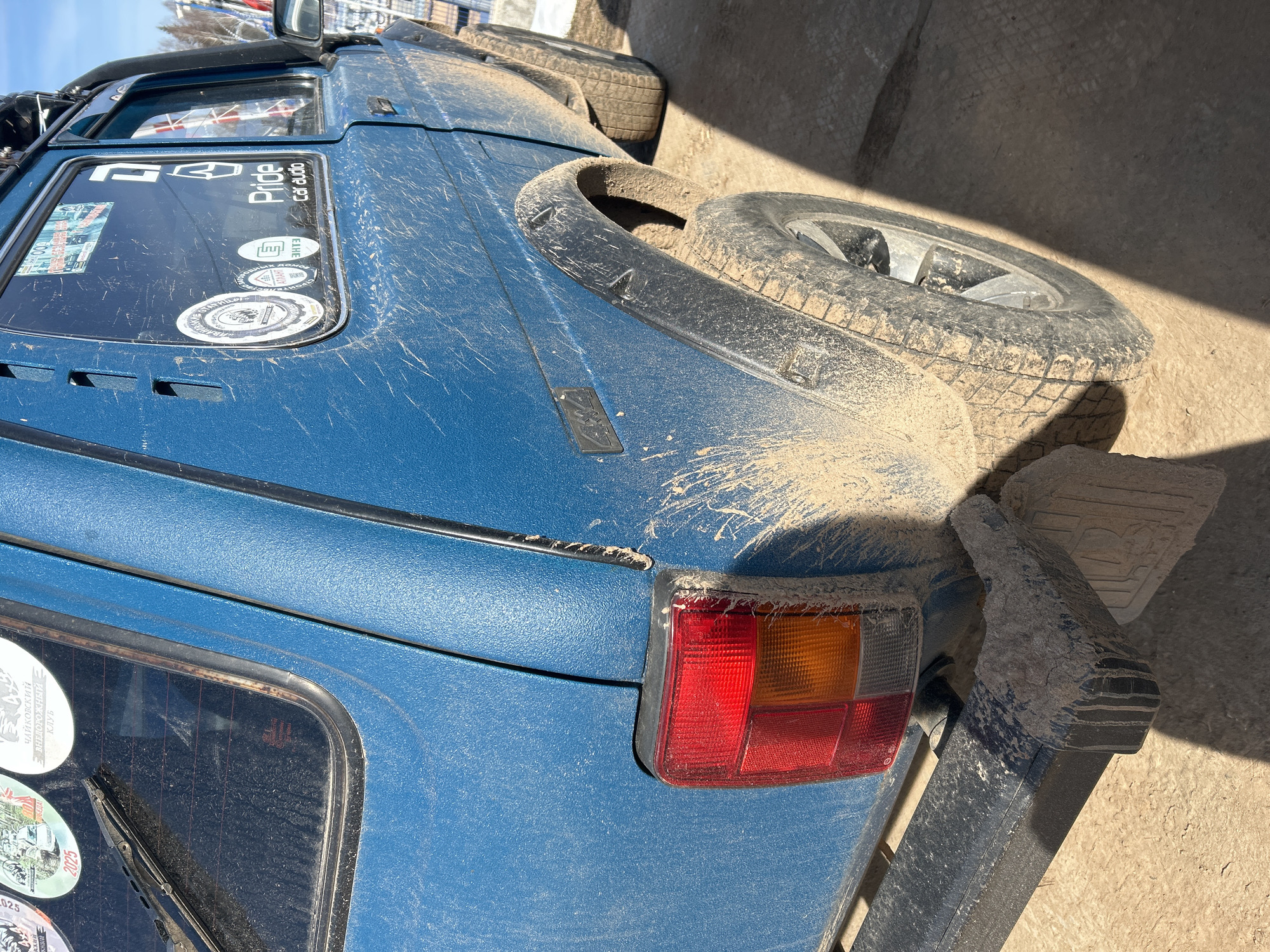 The image shows the rear of a blue, off-road vehicle covered in mud. The vehicle has a spare tire mounted on the hood. There are several stickers on the rear windows of the vehicle. The vehicle is parked on what appears to be a concrete surface. The photograph was taken in bright sunlight. Considering the window sticker mentioning "Чайковский внедорожный клуб", the image is most likely taken in Russia, possibly in the general area of Ural'skoe, Russia.