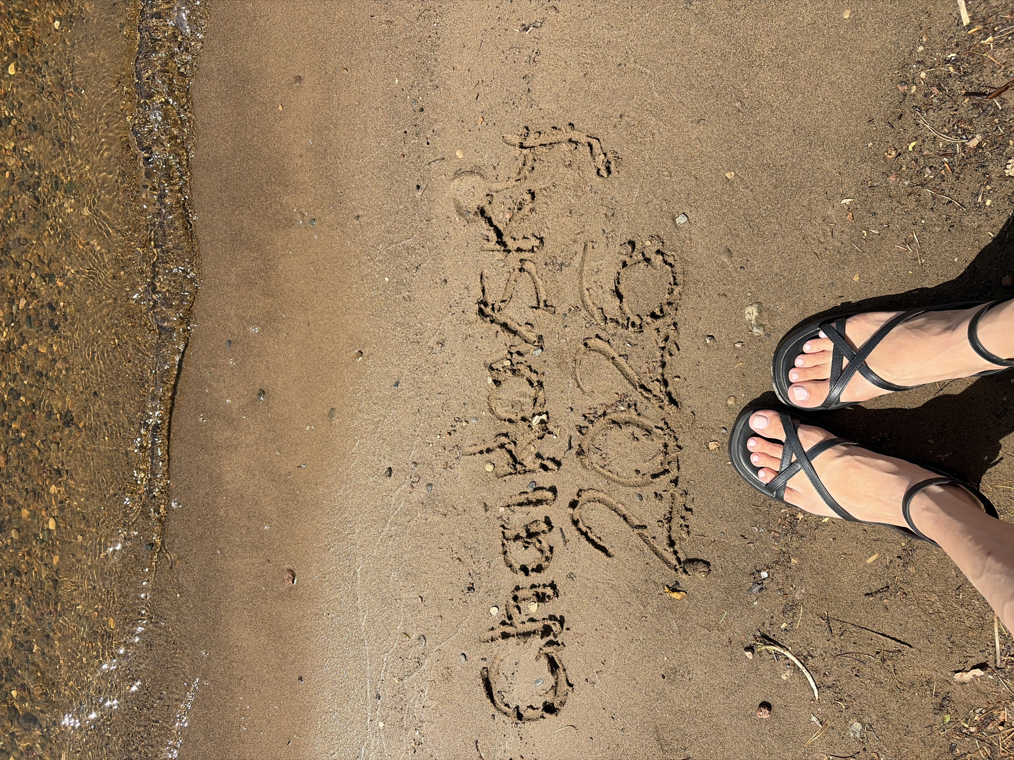 An overhead shot captures a sunny scene on a sandy shoreline, with clear, shallow water on the left and a person's feet on the right. The sand is light brown and appears damp near the water's edge. Visible in the sand, etched in cursive, is the text "Chaikovsky 2024," directly referencing the location context of Chaikovsky, Russia. The water, featuring small pebbles at the bottom, sparkles with sunlight, indicating a bright day. Distinct shadows cast by the person's feet and sandals further suggest strong sunlight, likely during midday or afternoon. The person is wearing black, strappy sandals, and their toenails are painted with a light pink polish. The overall scene depicts a peaceful moment by the water, possibly during a leisure activity or vacation, with the written message serving as a personal marker of the time and place.