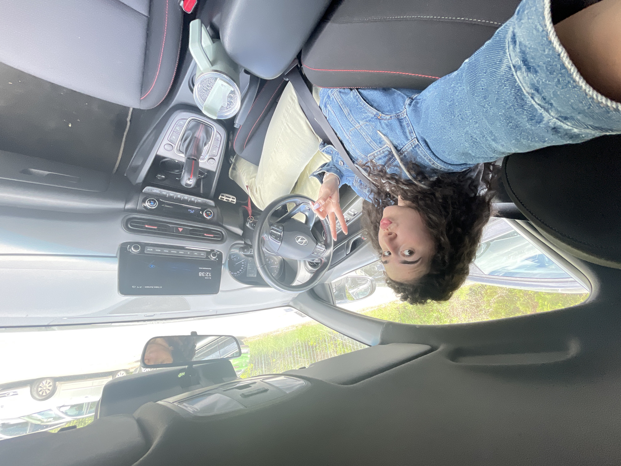 A person is sitting in the driver's seat of a car, oriented upside down in the image. The person has curly hair and is wearing a denim jacket. They are holding up a peace sign with their hand. The car's dashboard, steering wheel, and center console are visible. A drink cup is in the center console. Through the windshield, foliage and other cars are visible. The time on the car's dashboard is 12:38. There is no evidence in the image to determine a location in the city of Sofia, Bulgaria.