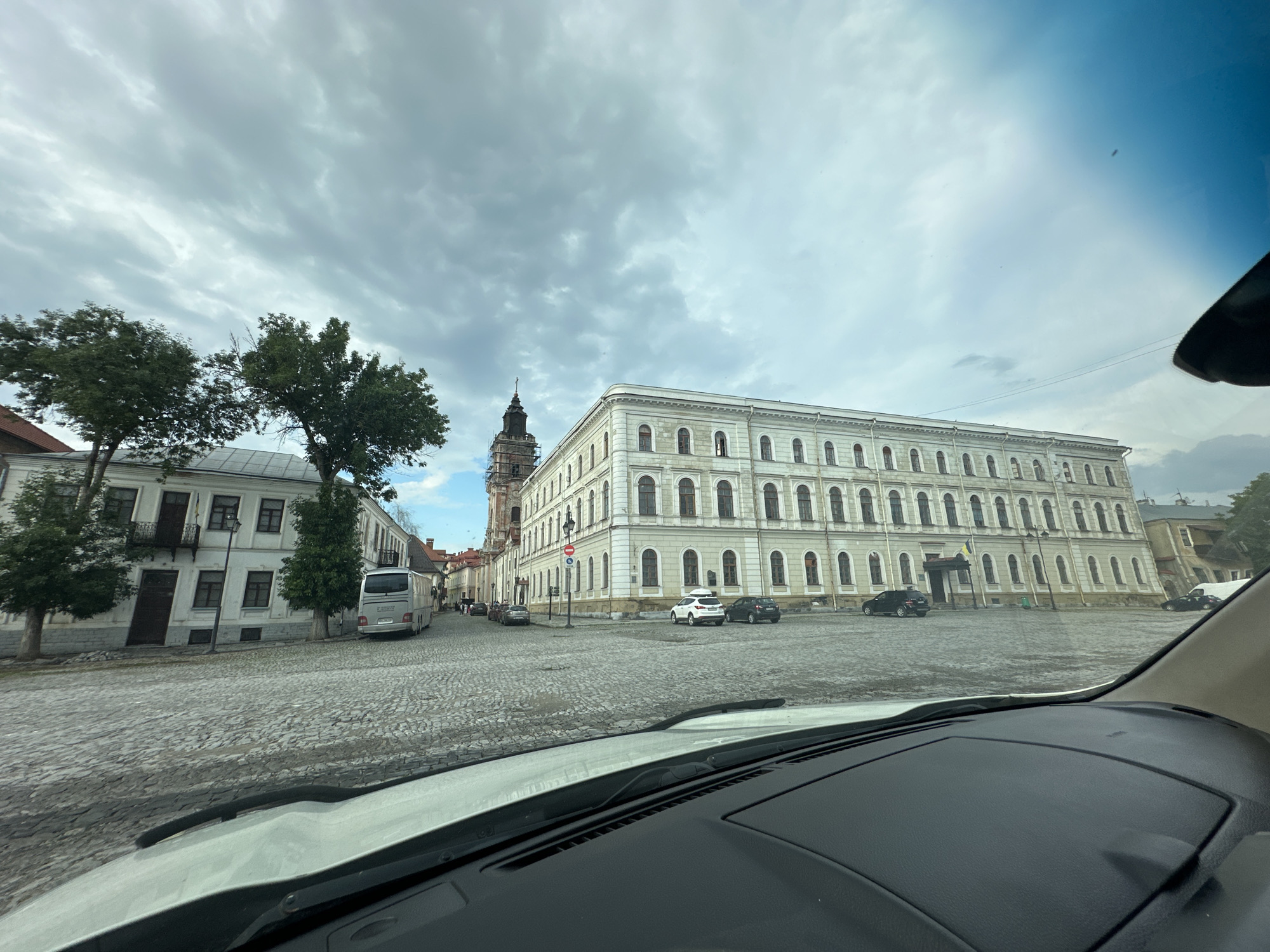 This media file captures a view from inside a vehicle, looking out onto a cobblestone street and a grand, historic building in Kamianets-Podilskyi, Ukraine. The building, characterized by its numerous arched windows and neoclassical architecture, dominates the right side of the frame. To the left, a narrower, older building with multiple stories and balconies stands. A tall, dark tower with a cross on top is visible between the two buildings, adding to the historical ambiance.
The street is paved with uneven cobblestones, suggesting an older urban area. Several cars are parked and driving on the street, including a white bus and a white SUV. The sky is overcast with dramatic, grey clouds, indicating either early morning or a storm approaching. The overall mood is subdued, reflecting the weather. No people are clearly visible, and the focus is on the architecture and the streetscape. The time of day appears to be daytime, given the ambient light, but the cloudy sky makes it difficult to be precise. There is no visible text in the image.