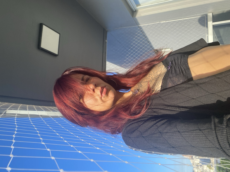 A woman with red hair is captured from a low angle, partially obscuring her face with shadows. She wears a black top, and visible ink adorns her right arm. A clear, protective net stretches across the foreground, with a bright blue sky visible through the gaps. In the background, a grey wall features a small, square frame. A cityscape is faintly visible in the distance, hinting at an elevated setting.