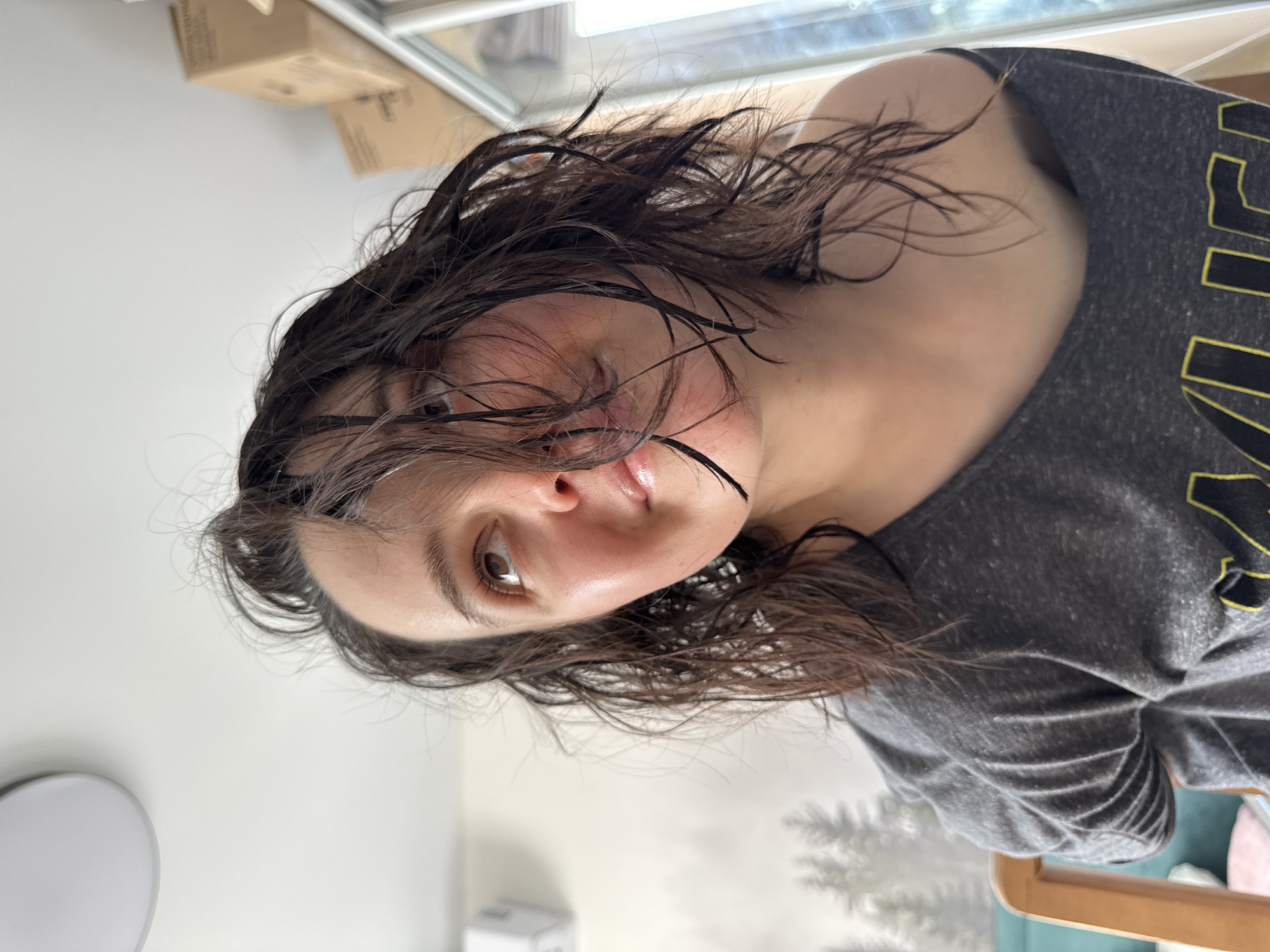 An adult female is depicted in a medium-close-up shot, looking upwards and to the right. She has dark, wet, disheveled hair, with strands falling across her forehead and face. Her skin appears fair, with a reddish flush on her cheeks. Her lips are slightly parted, exhibiting a glossy appearance. She is wearing a dark grey top or t-shirt, possibly off-the-shoulder, with a yellow design partially visible near the bottom edge of the frame.
The background on the left side features a white wall with a grey-scale mural depicting silhouettes of evergreen trees, a white wall-mounted unit (possibly an air conditioner or speaker), and the top section of a light-colored wooden chair. Behind the chair, a fragment of turquoise upholstery is visible. On the right side of the background, several plain cardboard boxes are stacked against a white wall, adjacent to a vertical window or glass door frame. A white circular ceiling light fixture is partially visible in the upper left corner of the frame. The specific location within Izhevsk, Russia, cannot be determined from the visual cues present in the image.