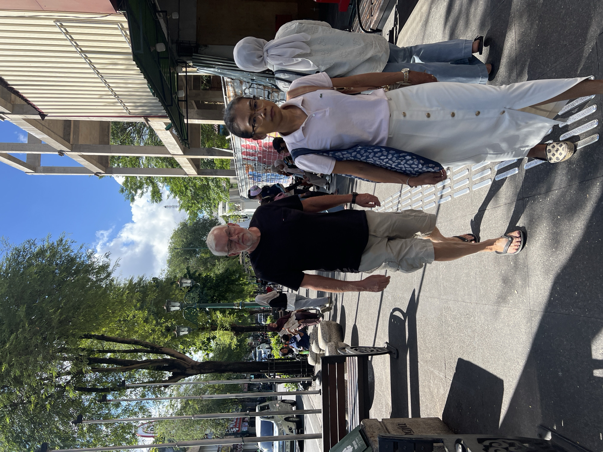 This is a medium shot photograph taken on a sunny day on a sidewalk in Yogyakarta, Indonesia. The foreground features a man with glasses, wearing a black t-shirt and light-colored cargo shorts, walking towards the camera. Beside him and slightly ahead, a woman with glasses is walking, wearing a pink polo shirt and a long white skirt. She is carrying a blue and white patterned bag over her shoulder. Behind them, a woman wearing a light blue hijab and a light blue top is walking away from the camera. In the background, several other people are seated on a bench, and further back, trees and buildings line the street. A white van is visible on the road to the left. The sidewalk is paved with square tiles and has tactile paving strips. The environment appears to be a busy urban street.