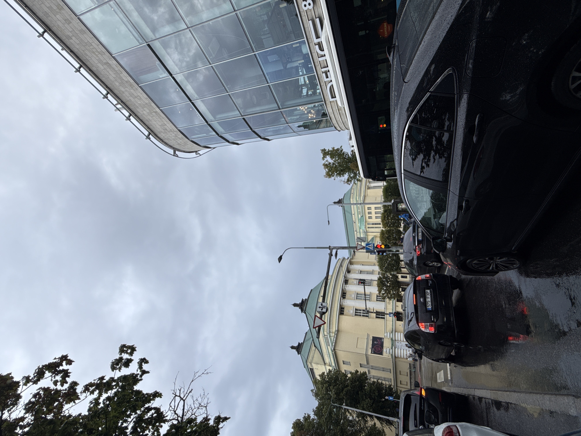 This is a street-level, medium-shot photograph taken in Tallinn, Estonia. The scene depicts a traffic intersection on a cloudy day, with wet pavement indicating recent rain. In the foreground, several cars are stopped, including a black sedan on the right and a black hatchback in the center with license plate "575 FXP". To the left, a white car is partially visible.
Dominating the background are two prominent buildings. On the left is a classical building with a pale yellow facade, white columns, and a green roof. It appears to be an institutional or cultural building, possibly a theater or opera house, with signage visible on its lower section. To the right of this building, a more modern structure with a curved glass facade reflects the overcast sky and surrounding environment. The words "PEUGEOT" and "8" are visible on the modern building's exterior.
A red traffic light is visible on a traffic signal pole in the center-right of the image, and a pedestrian crossing sign is also present. Trees with green foliage line the sidewalks. A dark-colored bus is seen stopped at the intersection behind the black hatchback. The overall atmosphere is urban and slightly somber due to the cloudy weather.