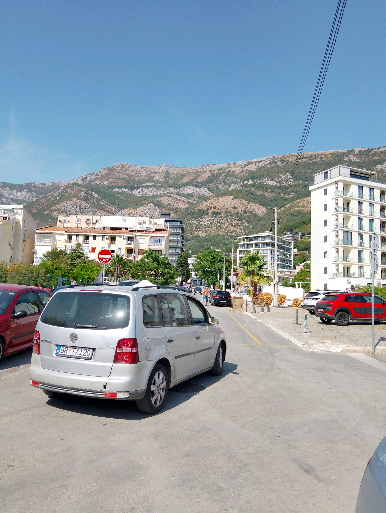 In Sutomore, Montenegro, the image captures a sunlit urban scene with a clear blue sky above rolling, verdant mountains in the background. A silver Volkswagen Touran is parked on a paved surface, its license plate reading "BR TX 120." To the left, a red car is partially visible. In the mid-ground, several buildings of varying architectural styles line a street, with palm trees and other vegetation adding to the Mediterranean ambiance. One building, a modern white structure with multiple balconies, stands out on the right. A red and white "no entry" sign is prominently displayed near a cluster of trees. The scene appears to be a typical street in a coastal town, possibly near a beach or promenade, with evidence of vehicular traffic and pedestrian activity further down the road.
