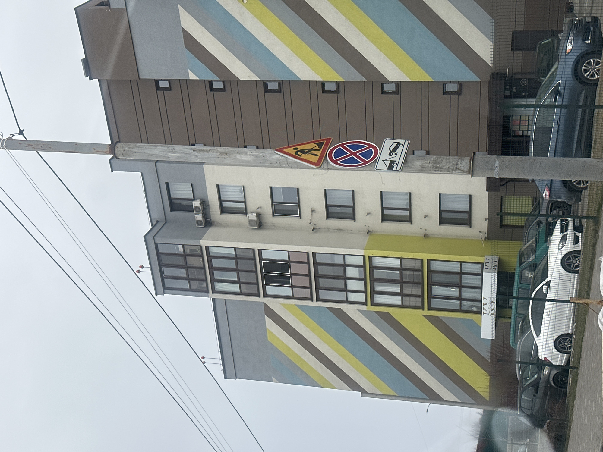 The image shows a street scene in Kyiv, Ukraine. The view is looking up at a multi-story apartment building with a modern design. The building's facade features a pattern of slanted stripes in light blue, yellow, white, and gray. Three road signs are attached to the building. Cars are parked along the street adjacent to the building. Overhead wires are visible in the sky. The sky is overcast.