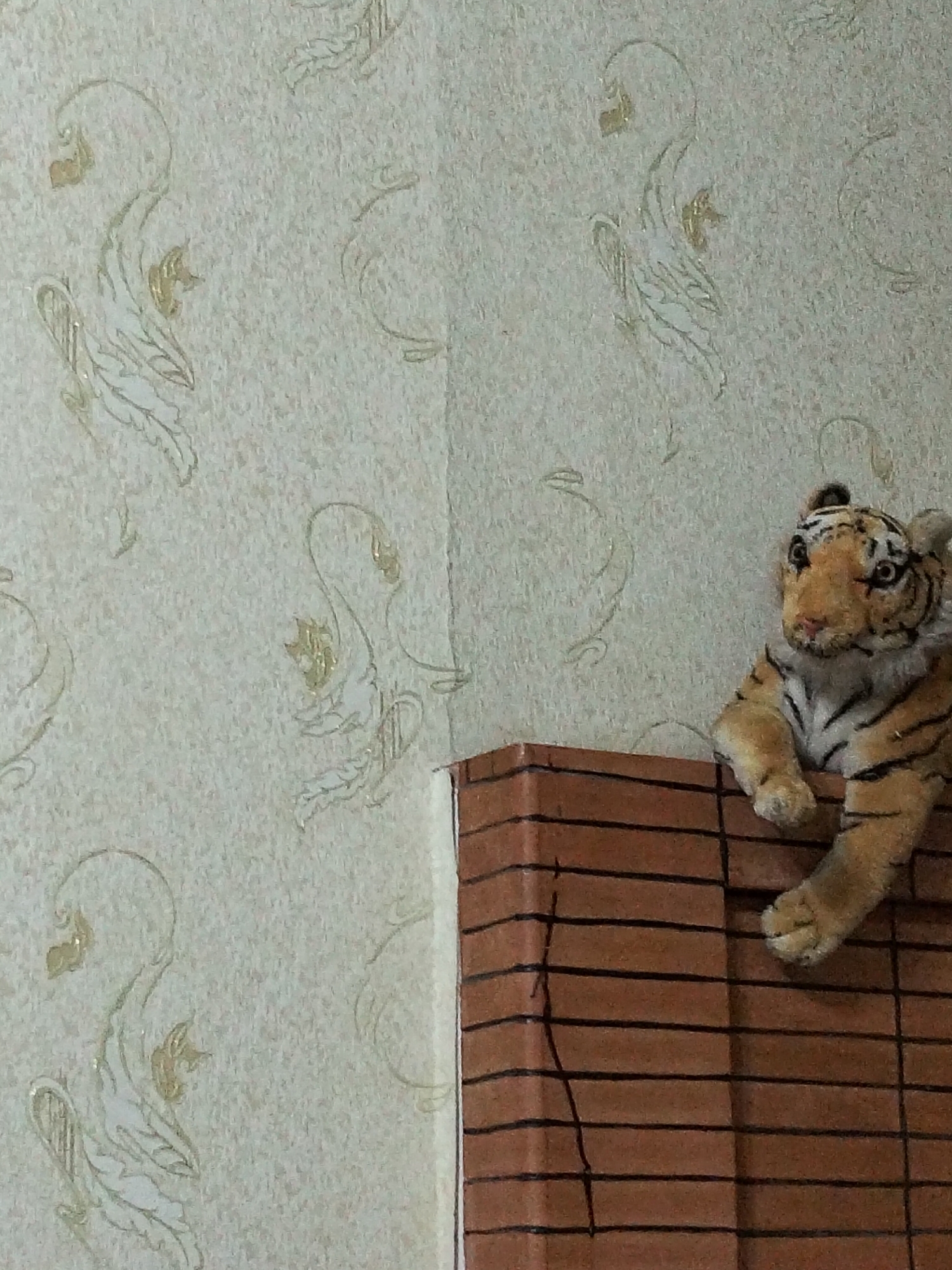 A plush toy tiger with orange fur and black stripes sits on a brown, brick-patterned surface. The tiger is positioned on the right side of the image, with its front paws resting on the surface. The background is a textured wallpaper with a repeating pattern of white and gold swan-like figures. The lighting suggests an indoor setting. The location in Kogon City, Uzbekistan, is not visually determinable from the image.