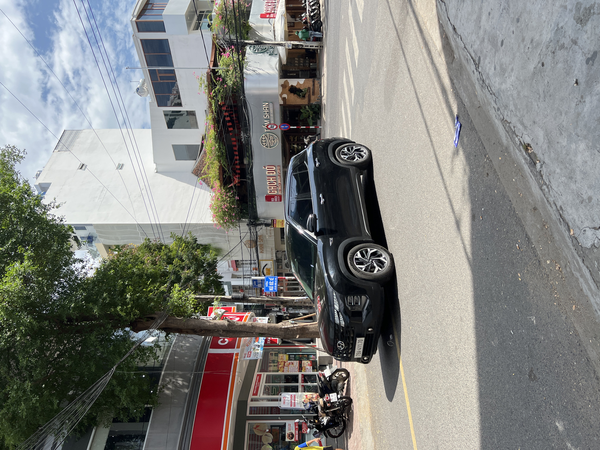 The image depicts a street scene in Nha Trang, Vietnam, under a partly cloudy sky. In the foreground, a black Hyundai Creta SUV is positioned on the right side of a road, facing towards the left. The vehicle's license plate reads "79A-411.70". To the left of the SUV, a portion of the sidewalk is visible, with two motorcycles parked. A person wearing a yellow shirt and blue shorts stands near the motorcycles.
The background features commercial buildings. On the left, a red and white building with the "CIRCLE K" logo is prominent. Further back, a white building with multiple floors and balconies is visible, alongside another building with a facade adorned with pink flowers and red banners. Signs on the buildings indicate businesses such as "GACH DO RED BEACH BAKERY & COFFEE" and "TÂM SHAN". Electrical wires crisscross above the street. The road surface is asphalt, with yellow lane markings and a pedestrian crosswalk in front of the SUV. Shadows are cast across the road and sidewalk, indicating it is daytime. Litter is present on the road and sidewalk.