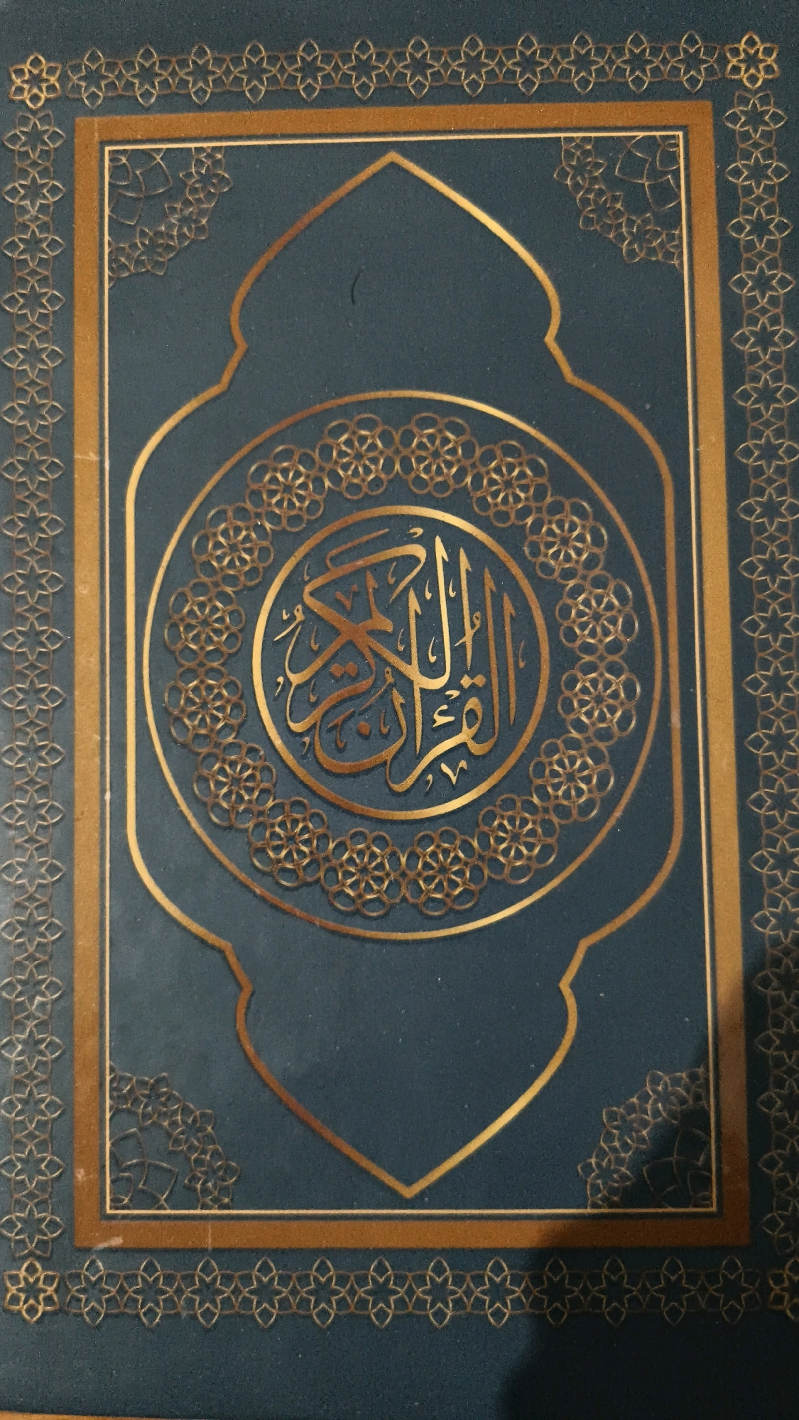 The image shows the cover of a book, likely a religious text, with intricate gold geometric patterns and Arabic calligraphy on a dark teal background. The cover features a large central circular motif containing the calligraphy, surrounded by decorative borders. The overall design is ornate and traditional. The image does not contain any information about the city of Poonch, India, nor does it depict any people, actions, or interactions.