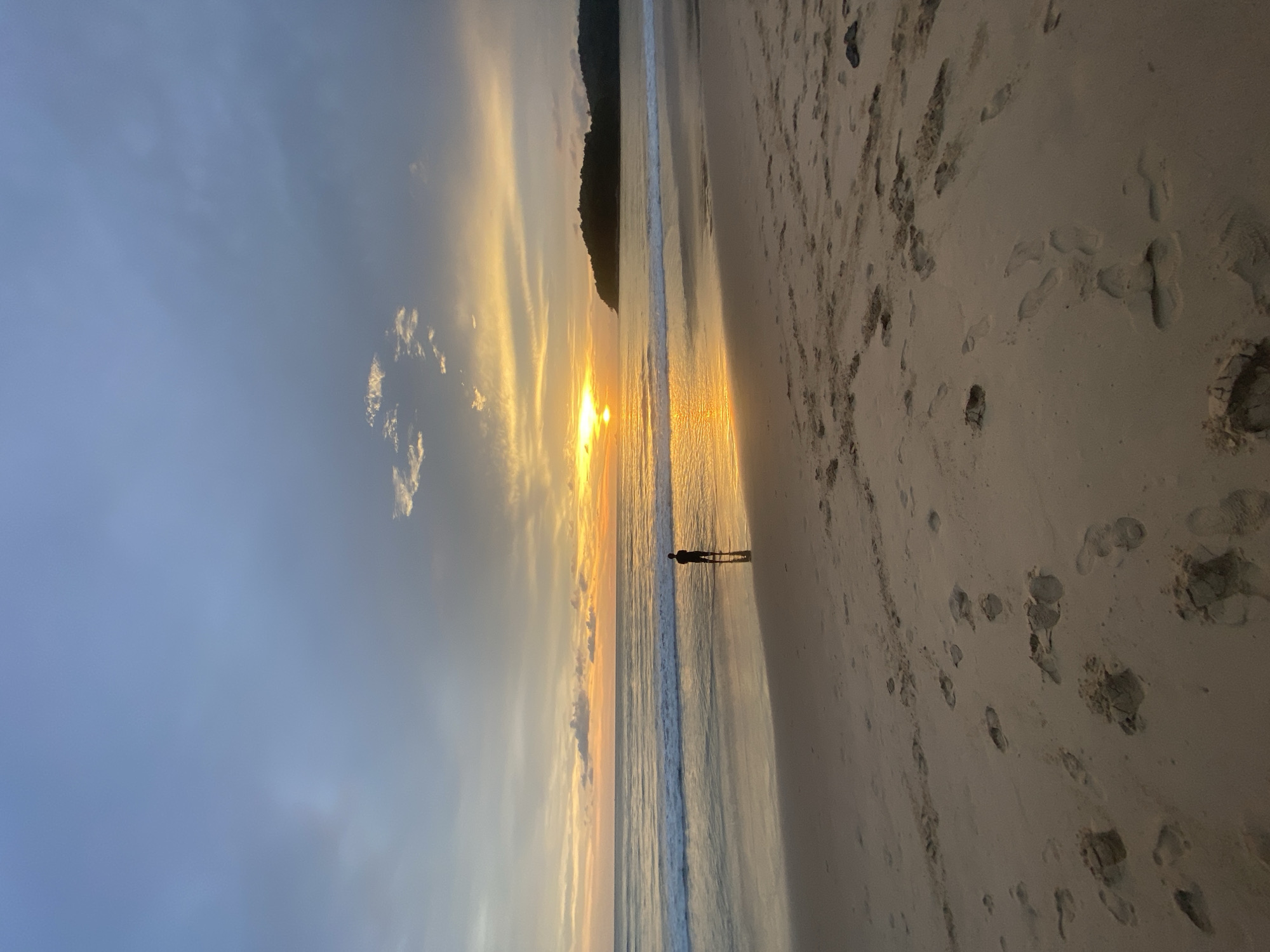 The image depicts a coastal scene at sunset, likely in Choeng Thale, Thailand given the query context. The sky transitions from a pale blue to orange and yellow hues as the sun sets on the horizon. Some scattered clouds are visible. A dark landmass is silhouetted in the distance.
The shoreline is sandy and wet, reflecting the sun's light. Footprints are visible on the sand. A lone person stands near the water's edge, casting a shadow on the wet sand. Small waves lap at the shoreline.