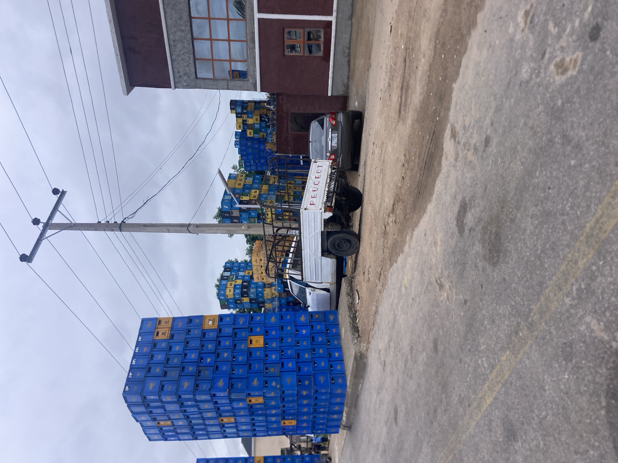 In Jalingo, Nigeria, a large stack of blue plastic crates is piled high on a concrete base. Adjacent to the crates, a white pickup truck with a cage-like back is parked. Behind the truck, a grey sedan is also parked. In the background, more stacks of crates, some yellow and some blue, are visible. A building with a maroon facade and large windows stands to the right of the scene, and a utility pole with overhead wires is positioned behind the stacks of crates. The ground in the foreground is an asphalt road with a yellow line.