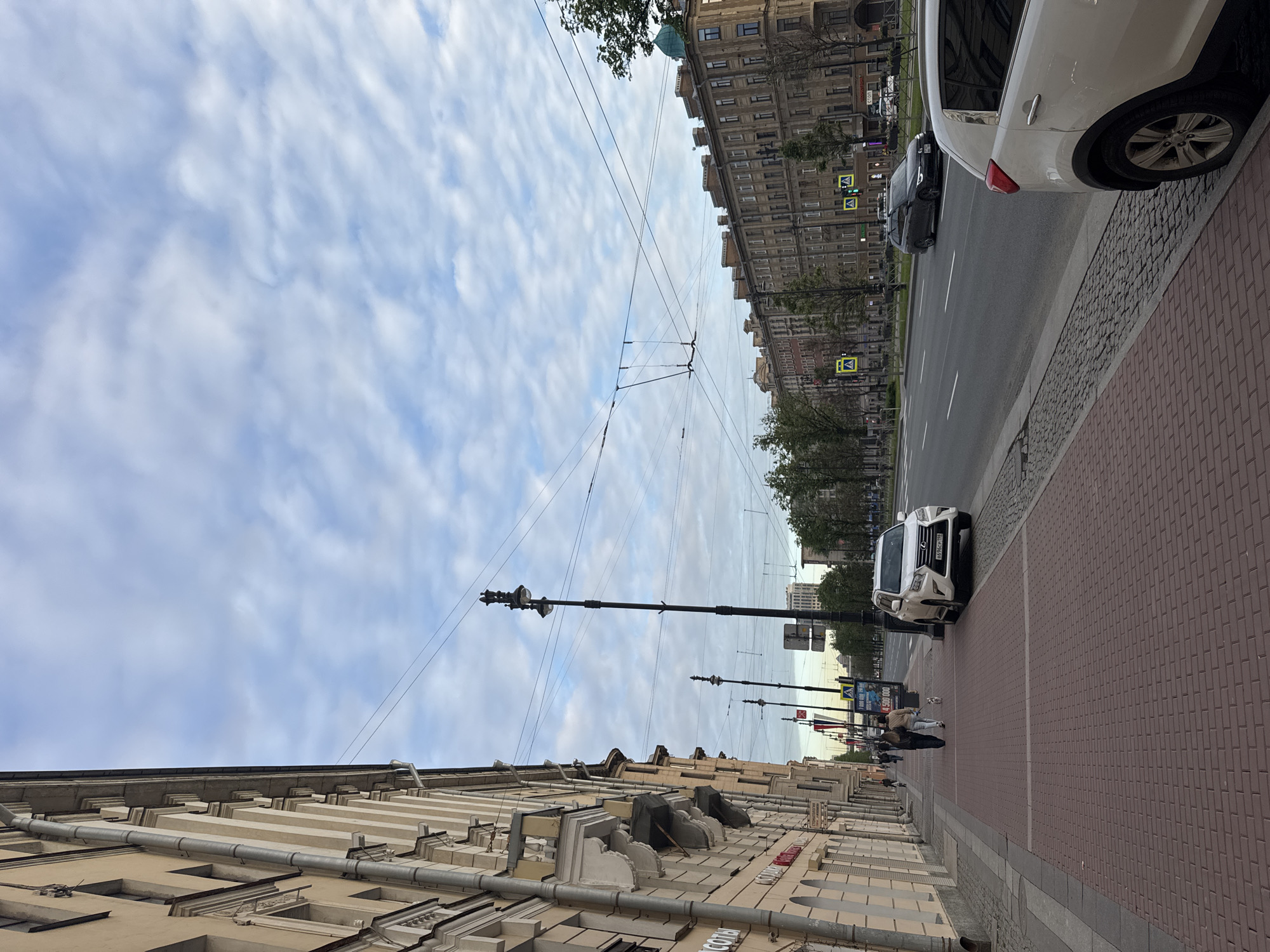The image captures a wide urban street scene in Saint Petersburg, Russia, on an overcast but bright day. The sky is filled with patchy white clouds against a light blue backdrop.
On the left side, a row of ornate, multi-story buildings with classical architectural details lines the street. These buildings are predominantly beige or light yellow, featuring prominent cornices, window frames, and downspouts. Some signs with Cyrillic text, such as "ЛОМБАРД" (Pawnshop) and parts of other words like "СКОБ," are visible on the building facades.
The right side of the image features a wide sidewalk paved with reddish-brown interlocking bricks, separated from the roadway by a grey curb. A white SUV, possibly a Lexus, is parked prominently on the edge of the sidewalk, displaying a Russian license plate "B599OP 799" (indicating Moscow region). Further down the street, another white car and a dark-colored car are visible on the road.
Pedestrians are few, with a couple of figures seen walking in the mid-ground, one pushing a stroller. Tall black streetlights with multi-pronged fixtures punctuate the sidewalk. Overhead wires, likely for trolleybuses or trams, crisscross the sky above the street. Green and yellow pedestrian crossing signs are visible in the distance, along with trees lining the street. A dark sign with "Ozon" and a large banner with "300 000" are discernible on buildings further down the street. The overall impression is a typical daytime streetscape in a historic European city.