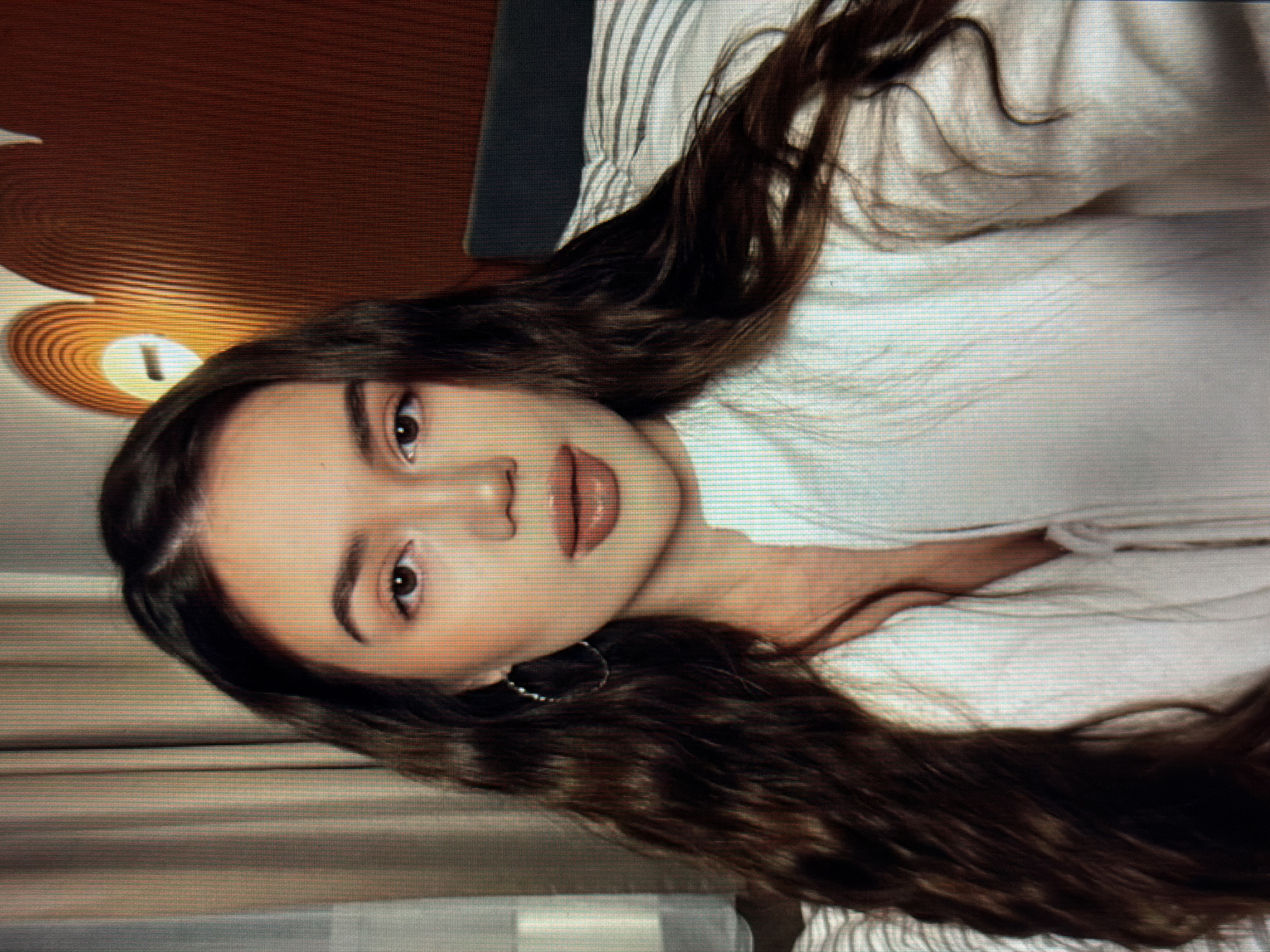 A medium shot shows a young woman with long, dark, wavy hair facing the camera. She is wearing a white collared shirt with a zipper. Her makeup is natural, with emphasis on her lips which are colored with a nude lipstick. She is wearing hoop earrings. The background is out of focus, but appears to be an indoor setting with curtains to the left and a textured wall with a curved light fixture to the right. The image quality is pixilated. The scene is likely captured in a residential setting within the city of Sirius, Russia.