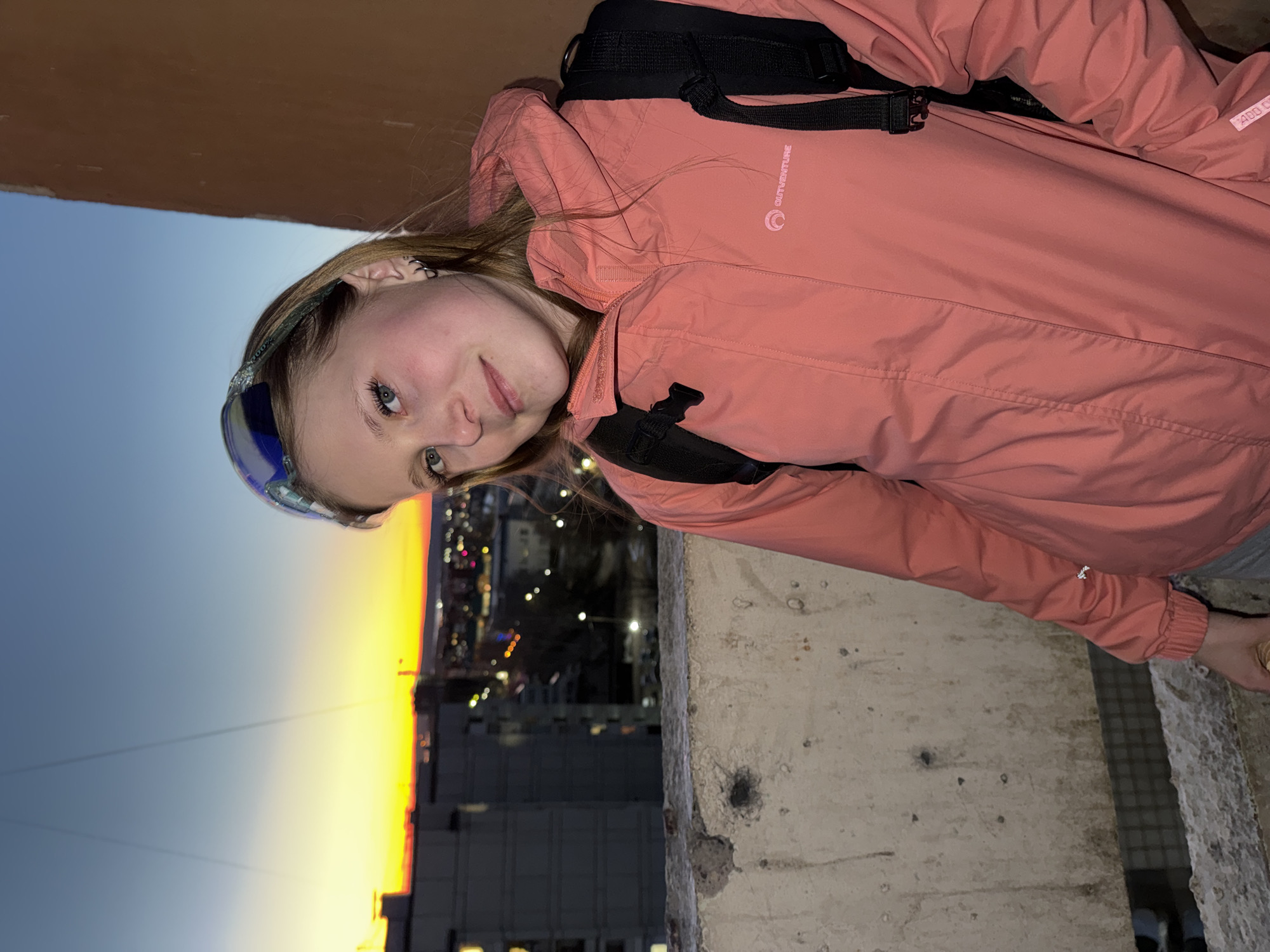 A medium, vertical shot presents a young woman in an orange jacket and black backpack, positioned against a backdrop of what appears to be a cityscape at dusk. Her face is turned towards the viewer with a slight smile. The woman is standing near a concrete structure.
In the background, the sky transitions from deep blue to yellow, indicative of a sunset or sunrise. Buildings are visible, some with lights on, suggesting it is evening. The location is Chaikovsky, Russia based on prior image analysis. The scene suggests an urban environment.