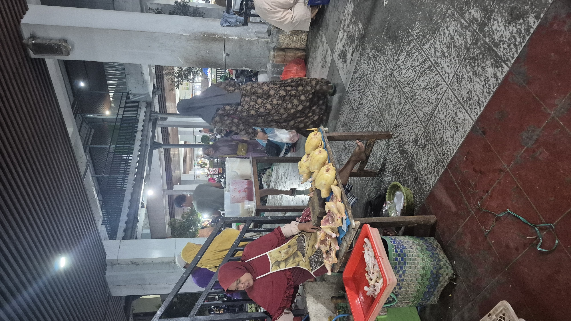 The image captures an outdoor market scene, likely at night or in low light, in Bojonegoro, Indonesia. In the foreground, a woman wearing a red hijab and a patterned apron sits at a wooden table, attending to raw chicken. Several whole chickens, some appearing to be prepped for sale, are displayed on the table. To the left of the table, a red plastic tray holds chicken parts, placed inside a green and blue woven basket. The ground is tiled, with a section of red tiles in the foreground and gray patterned tiles further back.
In the midground, several people are visible. To the left, a person in a yellow hijab stands near a staircase. Further back, a person in a green shirt is seen walking, and another individual in a brown hijab is standing. A woman in a dark floral dress walks away from the camera towards the right side of the frame. Various goods, including bags and baskets, are scattered around the market stalls. In the background, structures with slatted ceilings and railings suggest an elevated walkway or covered market area, with artificial lighting illuminating the space. The overall impression is a bustling, informal marketplace.