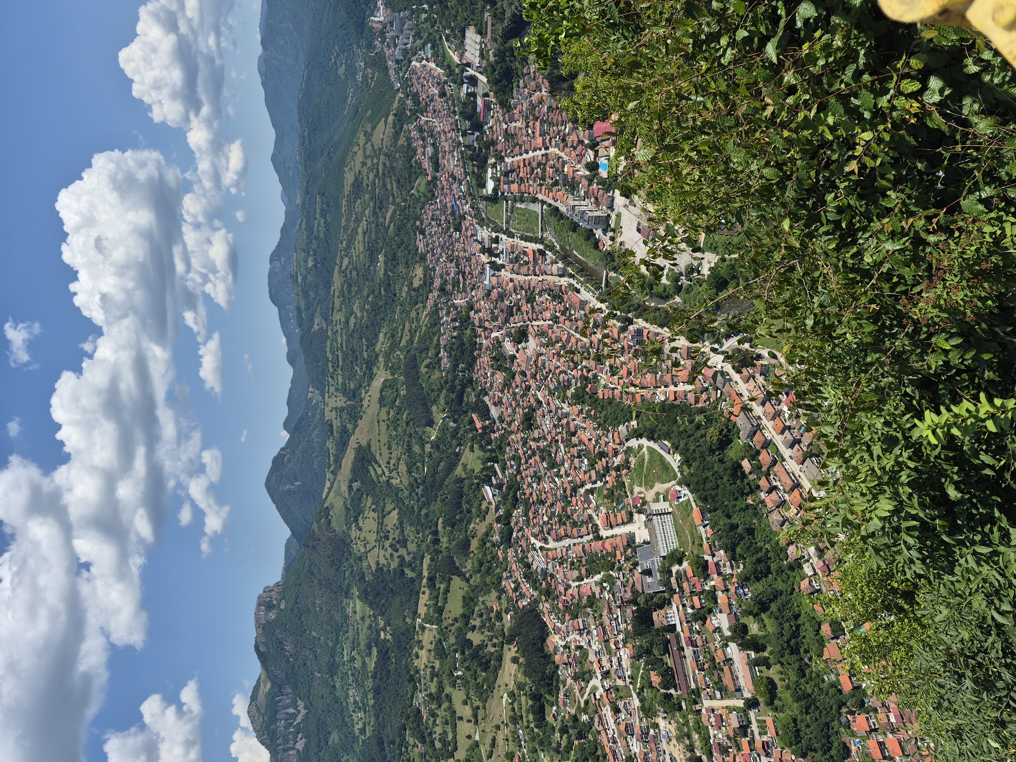 This is a high-angle, wide shot of the town of Teteven, Bulgaria, on a bright, sunny day with scattered white clouds in a blue sky. The town is nestled in a valley surrounded by lush green, rolling hills and distant mountains. The buildings in Teteven are predominantly red-tiled, closely packed, and appear to be residential and commercial structures. A river or stream runs through the middle of the town, with bridges crossing it. Several winding roads are visible, connecting different parts of the town and leading into the surrounding countryside. In the foreground, dense green foliage, likely bushes and trees, partially obscures the view of the town. The overall impression is of a picturesque and tranquil mountain town.