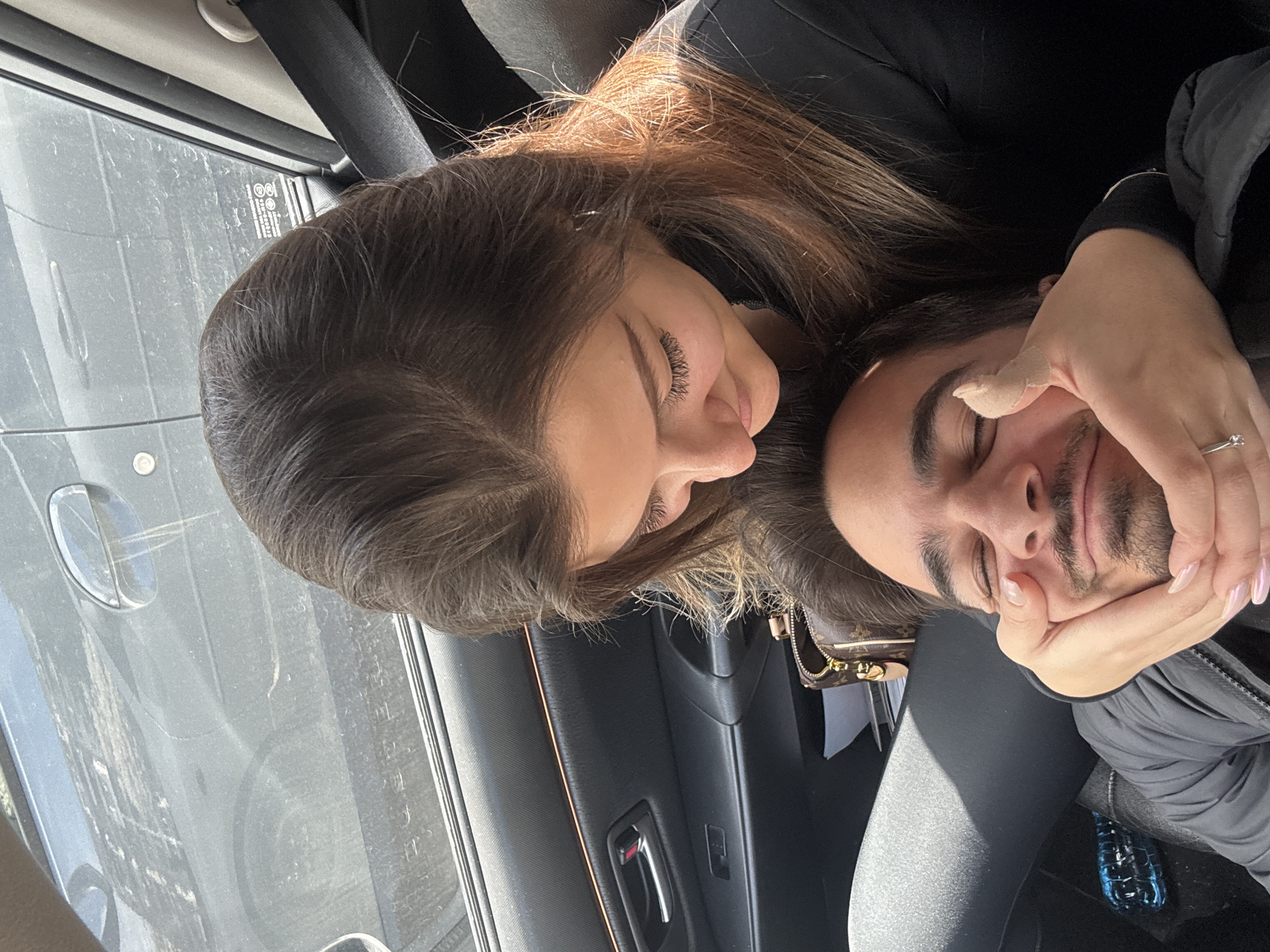 Two people are seated in a car. A woman with long brown hair is looking down at a man with dark hair and a beard. The man has his eyes closed and is resting his head in the woman's hand. The woman is wearing a ring on her left hand. The interior of the car is dark, with a gray door panel and black seats. The exterior of the car is visible through the window, showing a gray car and an indistinct background. There is no indication of the city or the country.