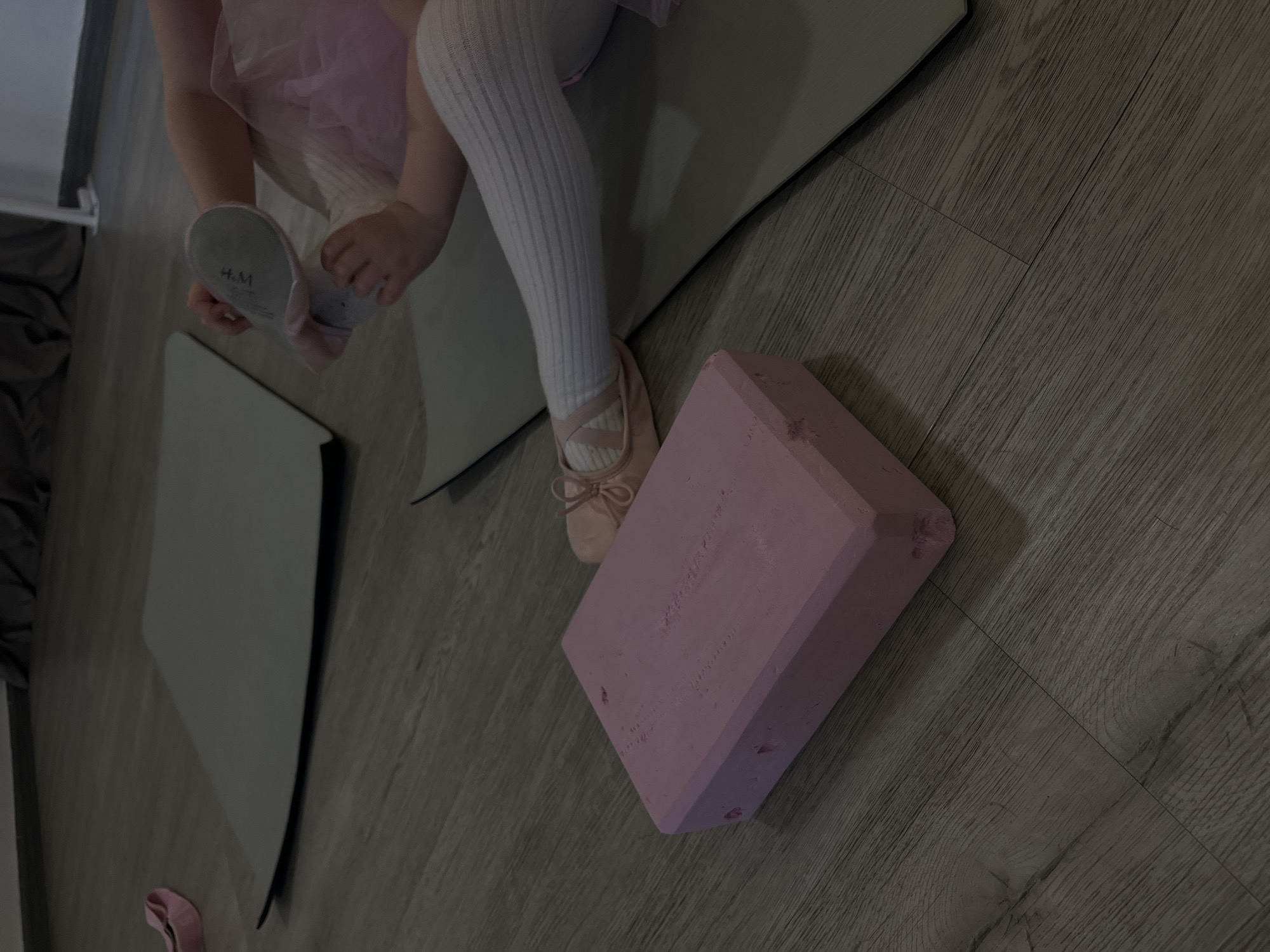 A dimly lit, angled shot reveals a child putting on a ballet shoe. The child is wearing white leggings and a pink tutu. One shoe is partially on the child's foot, revealing the "H&M" logo on the sole. The other foot is already adorned with a ballet shoe. The child is sitting on a grey yoga mat. A pink yoga block is situated on the wood-look floor. The location is indiscernible, with no clues pointing to Budva, Montenegro. The primary focus is on the child and ballet equipment.