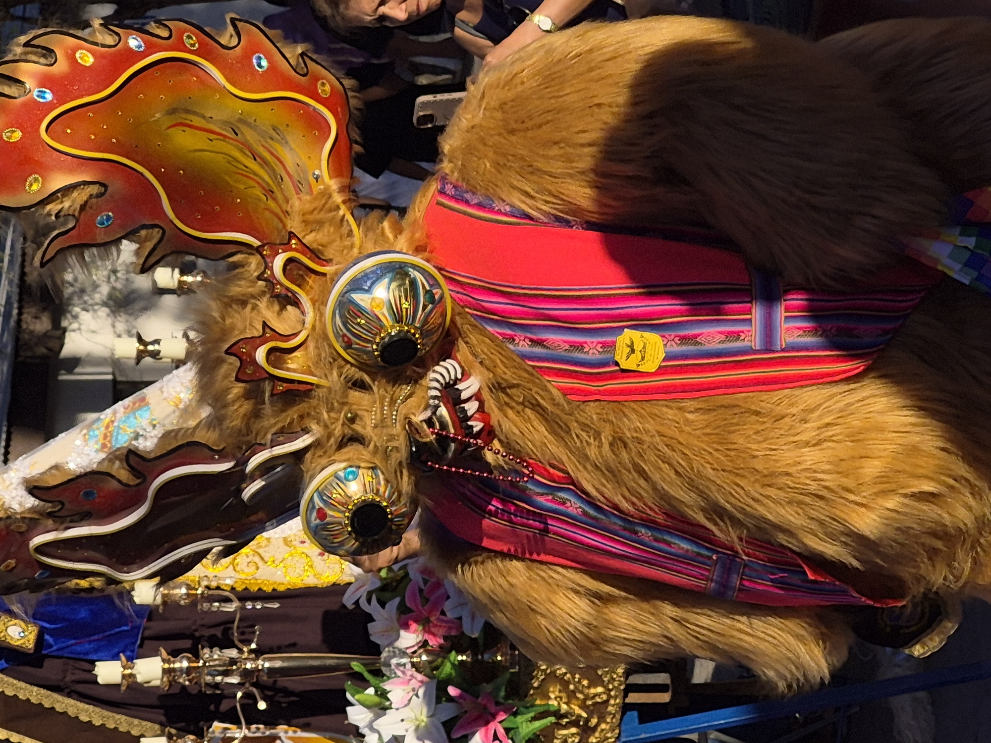 The image captures a person dressed in a detailed costume, likely for a festival or celebration, in Pozo Almonte, Chile. The costume features a furry, brown, bear-like body with a prominent, decorative mask for a head. The mask has large, elaborate, circular eyes with colorful, metallic-looking irises and dark pupils. Above the mask, the costume is adorned with large, flame-like headdresses or horns, colored in vibrant shades of red, yellow, and orange, decorated with jewel-like embellishments. The costume also includes a red sash with a colorful, striped pattern, possibly made of woven fabric, draped across the chest. A beaded necklace hangs around the neck area of the costume. In the background, partially visible, are elements of a festive or religious procession, including decorative candle holders with lit candles, flowers (white and pink lilies), and draped fabrics in dark blue and gold. To the right of the main subject, a woman with dark hair is partially visible, wearing a dark top and a watch on her left wrist. The setting appears to be outdoors, with a slight shadow cast on the costume, suggesting it is daytime.