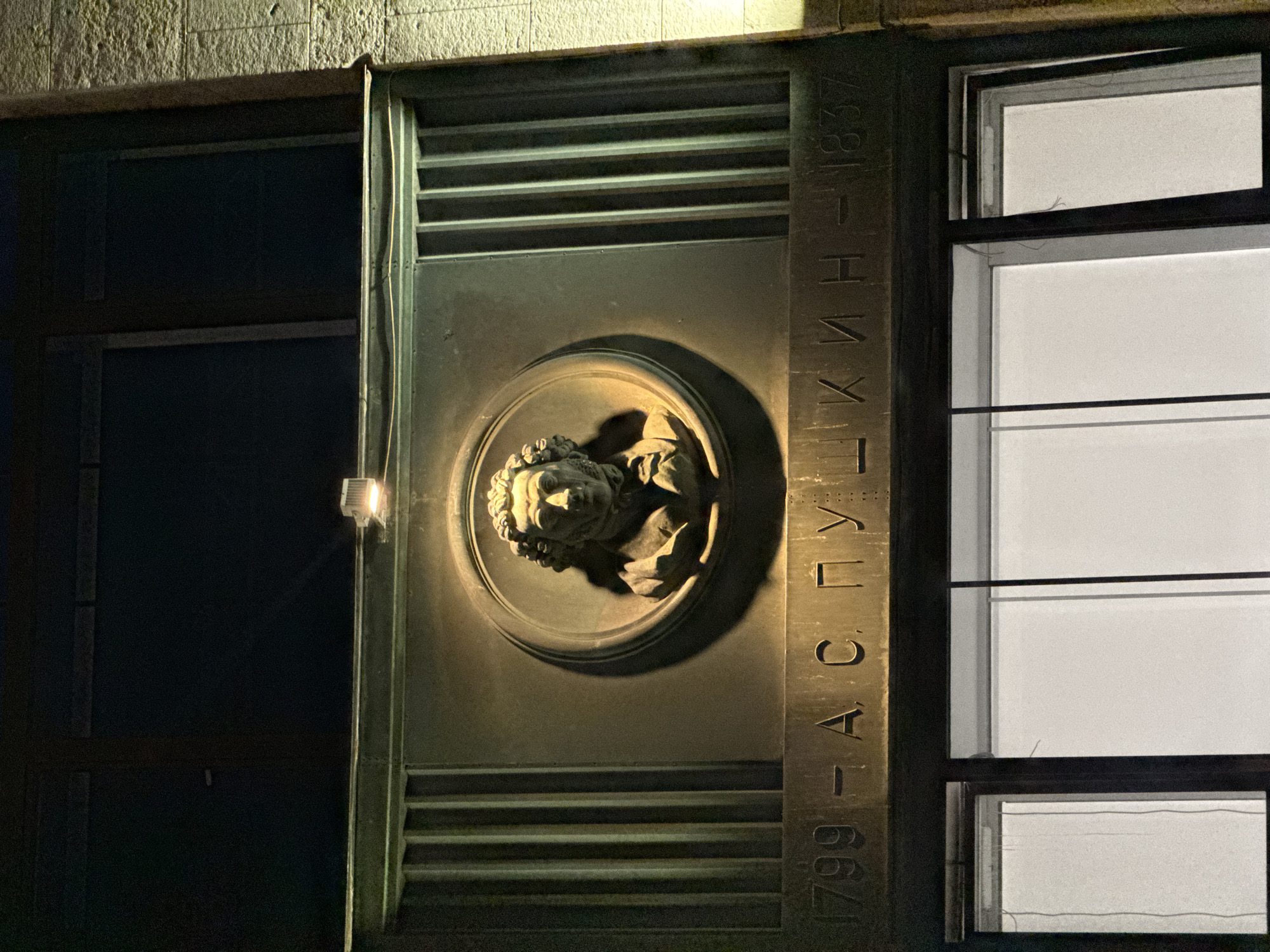 The image shows a relief bust of Alexander Pushkin, a prominent Russian poet, set within a circular frame on a building facade. The relief is mounted on a bronze plaque that also bears the dates "1799 - A. S. PUSHKIN - 1837" in raised lettering. The plaque is illuminated by a spotlight positioned above it. Below the plaque are windows, one of which is partially obscured by glare. The building appears to be constructed of stone and metal. The overall scene suggests a monument or memorial dedicated to Pushkin. This monument is located on the facade of the State Literary Museum in Moscow, Russia.