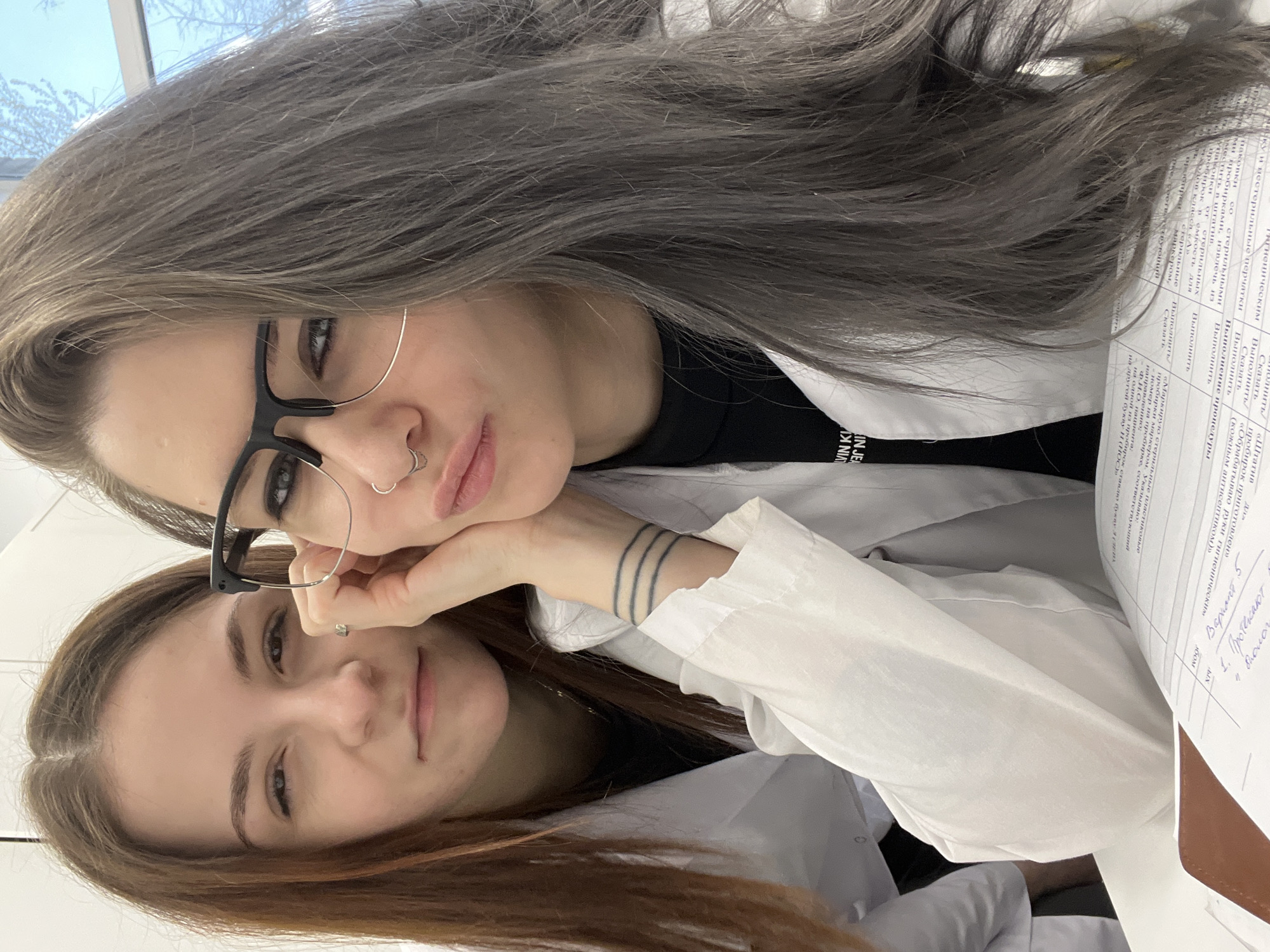Two young women wearing white lab coats are captured in a close-up, upward-angled selfie-style photograph, likely taken indoors in what appears to be a laboratory or classroom setting in Moscow, Russia.
The woman in the foreground has long, straight ash-brown or gray hair, black-framed glasses, and a nose piercing. She rests her chin on her hand, displaying a tattoo of three parallel lines on her left wrist. A black garment with partial English text "IN JEA" (likely part of "CALVIN JEAN") is visible under her lab coat. Her expression is calm with a slight pout.
Behind and slightly below her is another woman with long, reddish-brown hair, looking directly at the camera with a subtle smile.
The background shows a bright window with bare tree branches against a light blue sky, indicating daytime. To the right, several documents with handwritten and printed Cyrillic text are visible, consistent with the specified Moscow location context. Visible Cyrillic text includes headings and instructions such as "Гигиеническим", "Яку и нестерильные перчатки" (hygienic and non-sterile gloves), "Выполнить" (perform), "Сказать" (say), "Вариант 5" (Option 5), "1. Протекают", "Штатив" (stand), "пробирок" (test tubes), "стерильными" (sterile), "Маркирую стерильные пластиковые пробирки" (I mark sterile plastic test tubes), "Ф.И.О. пациента" (patient's full name), "направлению" (direction), and "букву Н (НОC)" (letter H (NOS)). The overall mood of the image is casual and relaxed.