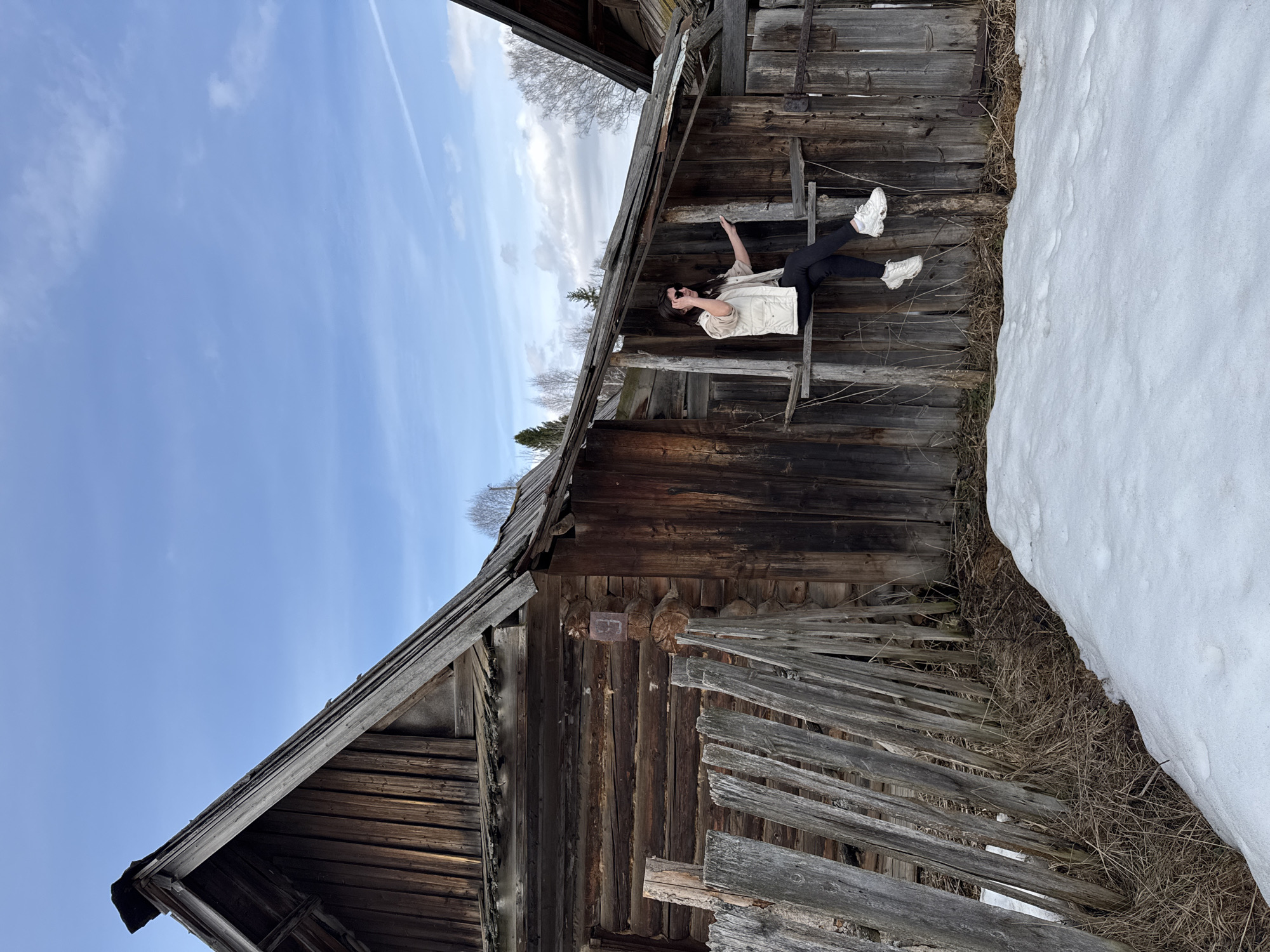 The image depicts a person sitting on a dilapidated wooden structure, possibly an old barn or shed, in Вассята, Russia. The structure is constructed of weathered wood and features a partially collapsed fence. The person is wearing a light-colored jacket, dark pants, and white sneakers. A patch of snow is visible next to the structure. The sky is blue with some clouds.