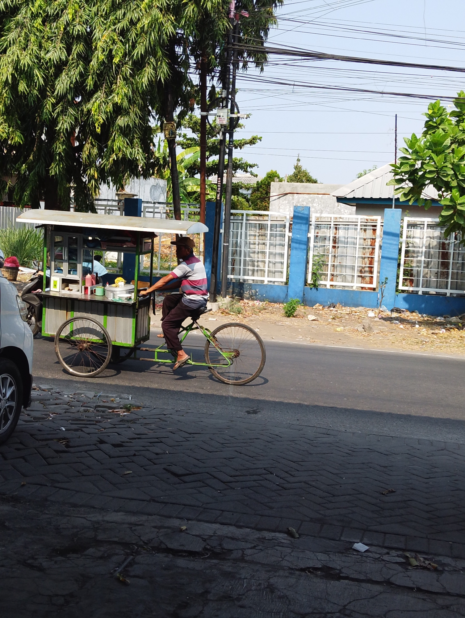 A man wearing a striped t-shirt and a brown hat rides a green bicycle with a cart attached. The cart is white on top and green below, with clear panels in the front and side displaying various food items and condiments. The bicycle is moving on a paved road, to the right of the frame. In the background, there is a blue and white building with barred windows, trees, and utility poles with many wires. The ground is paved with interlocking bricks, and there is some debris scattered on the side of the road. A white car is partially visible on the left side of the frame. The scene appears to be in Manaruwi, Indonesia.